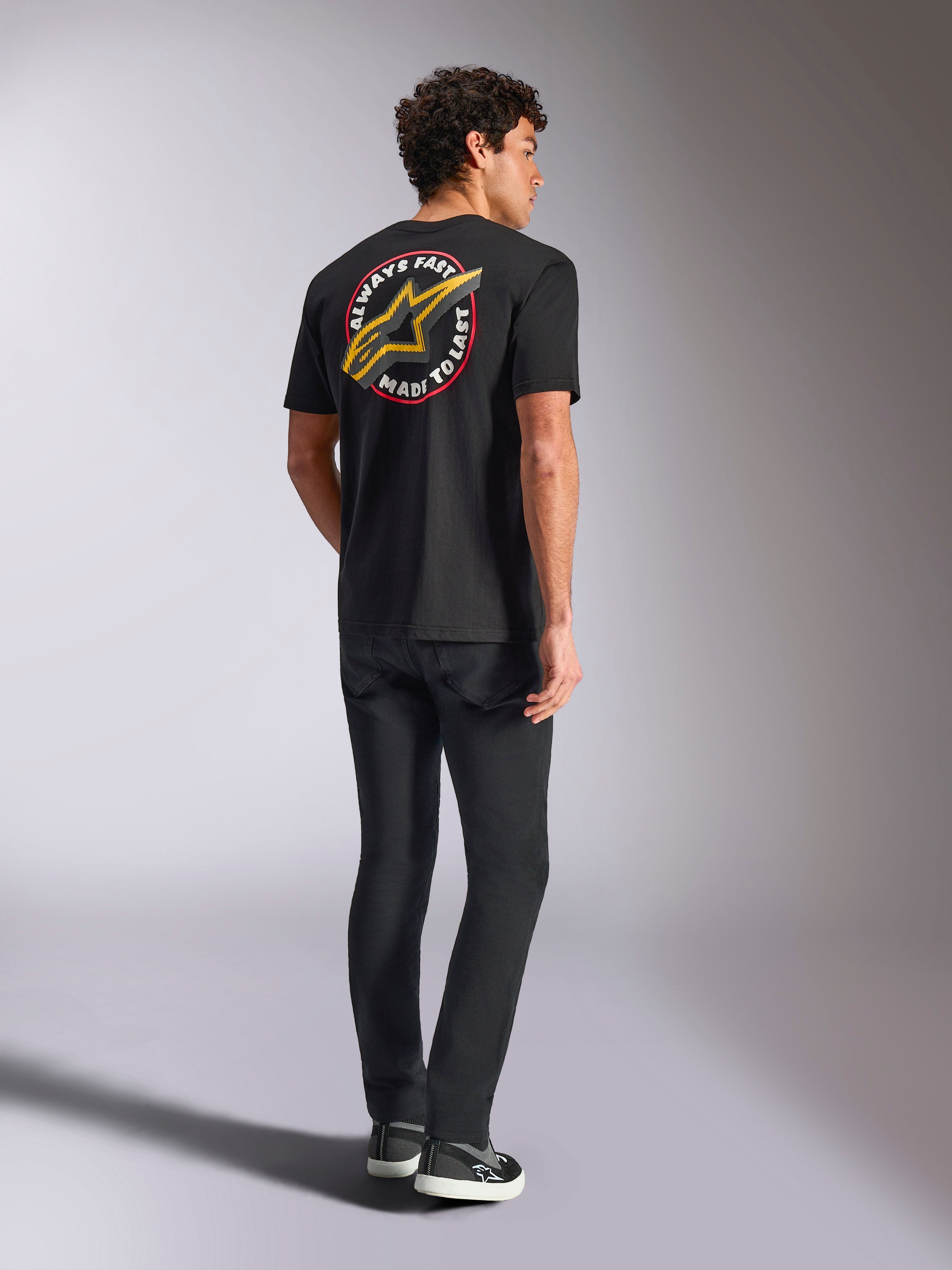 Alpinestars Aorbit CSF Tee, T-shirt de manga curta, preta, vista traseira com um logótipo Astars grande e amarelo e o gráfico circular 'Always Fast Made to Last', usada com calças de ganga pretas e sapatilhas casuais, estilo streetwear inspirado no desporto motorizado