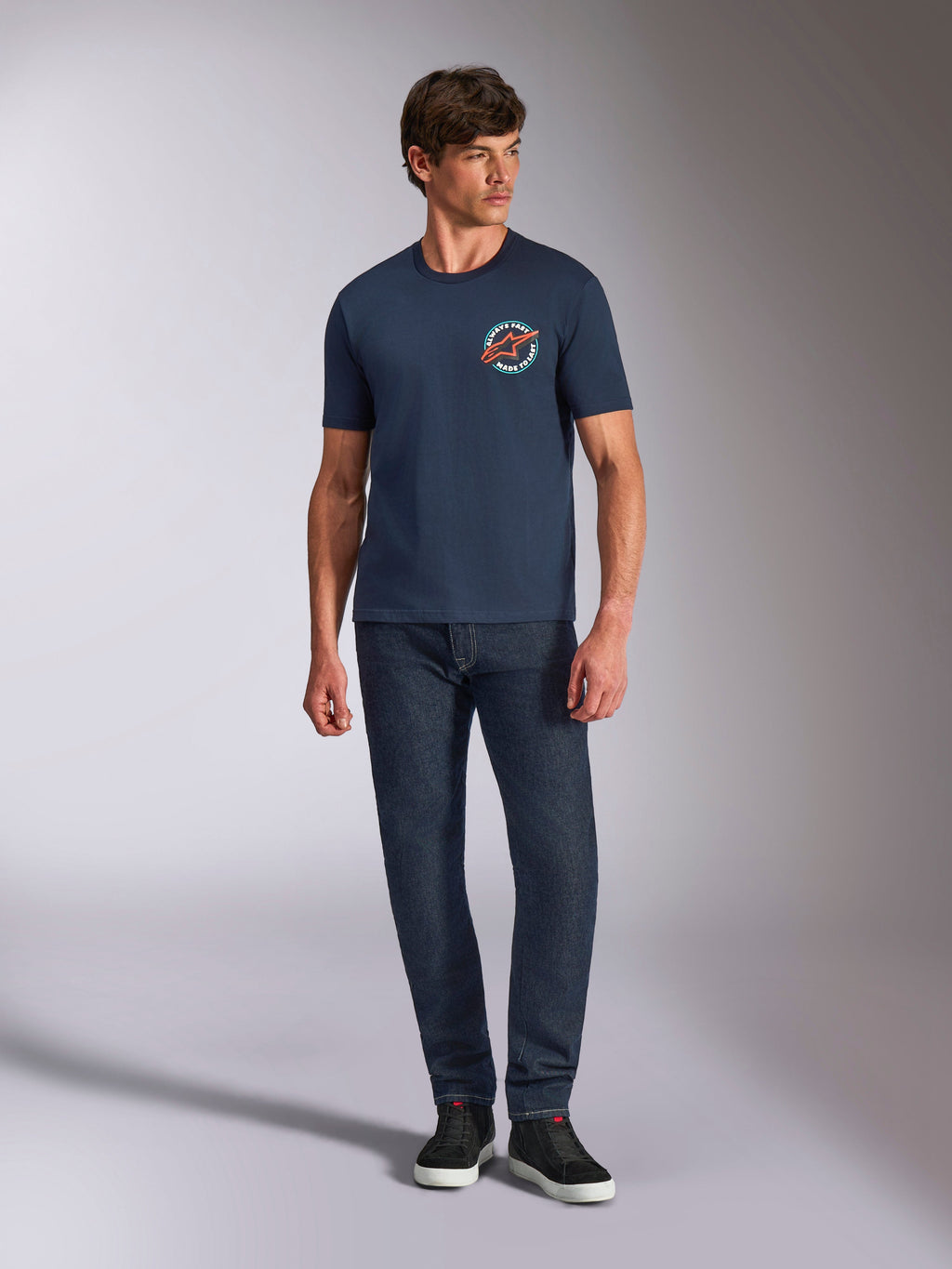 T-shirt Alpinestars Aorbit CSF, t-shirt de manga curta, azul marinho, gráfico circular no peito com o texto "Always Fast Made to Last", usada com calças de ganga escuras e sapatilhas, vestuário casual de estilo de vida motorsport