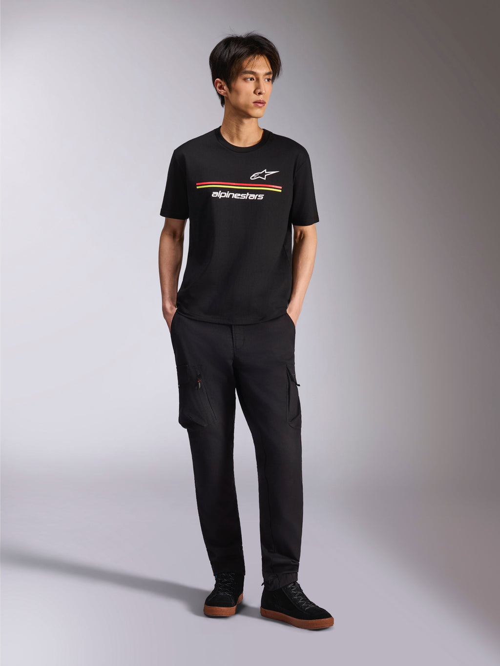 Modelo a vestir a T-shirt Alpinestars Lanes CSF, t-shirt de manga curta, preta, com riscas vermelhas e amarelas e logótipos Astars e Alpinestars, combinada com calças cargo pretas e ténis, streetwear casual de motorsport