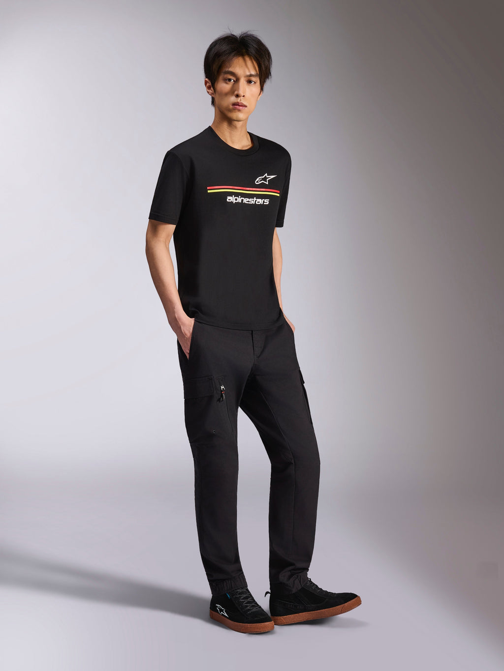 Alpinestars Lanes CSF Tee, T-shirt de manga curta, preta, logótipos Astars e Alpinestars com riscas multicolores no peito, vestida por um modelo com calças cargo pretas, streetwear casual de desportos motorizados