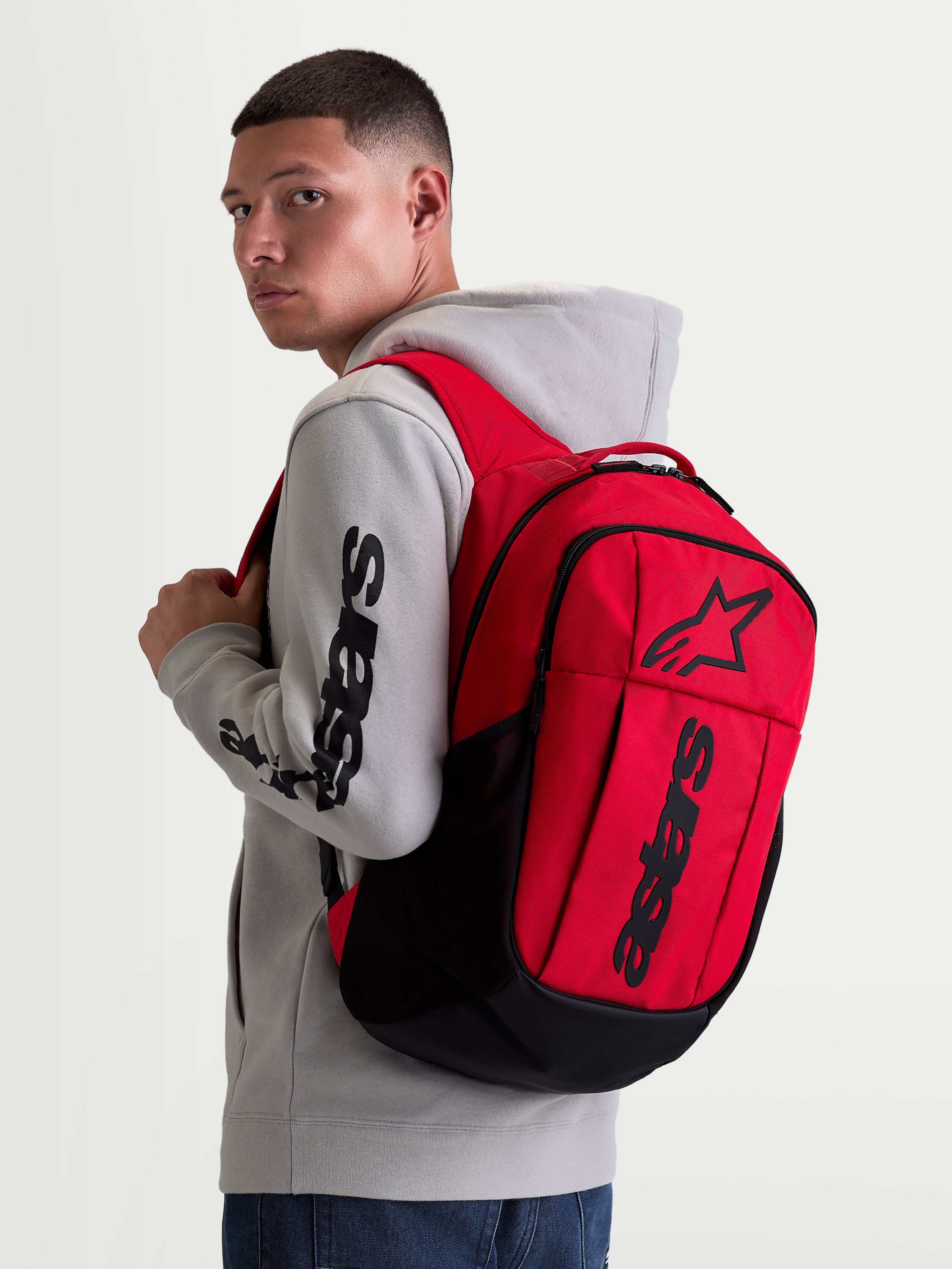Alpinestars GFX Boss, mochila urban sportswear de 20L, vermelha e preta, mostrada num modelo a usar um casaco com capuz cinzento. Inclui construção em poliéster 600D e logótipo Astars em relevo de silicone. Ideal para o dia a dia.