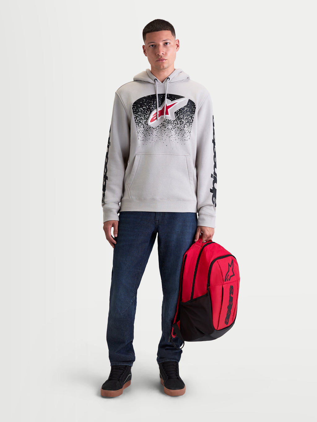 Mochila Alpinestars GFX Boss, mochila de dia para sportswear, vermelha e preta, capacidade de 20L, fabricada em poliéster 600D resistente com logótipo Astars em silicone, segurada por um modelo masculino para uso urbano casual.