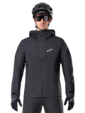 Alpinestars A-Dura Elite Waterproof Jacket, Black, Mountain Bike Jacket, High-collar design with hood, Front zipper with protective chest flap and white logo, Worn with black helmet and cycling gloves, Technical weather protection for trail riding