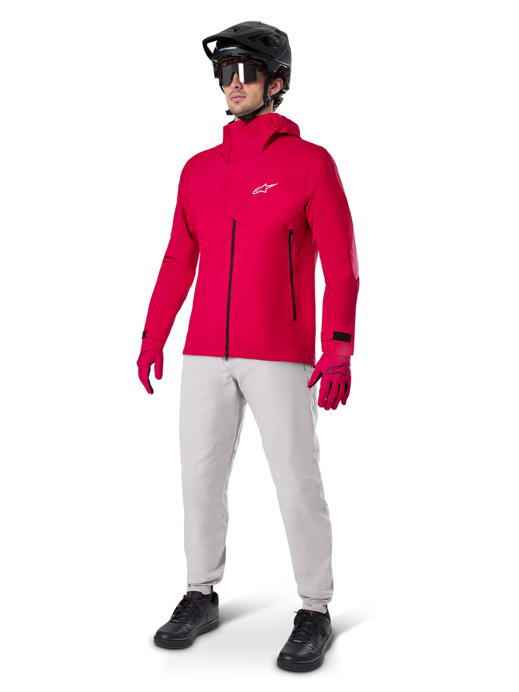 Ciclista masculino com o Casaco Impermeável Alpinestars A-Dura Elite em Vermelho Fluo, calças técnicas cinzento claro, luvas vermelhas, capacete de BTT preto com óculos e sapatos pretos, equipamento completo de alto desempenho para mountain biking