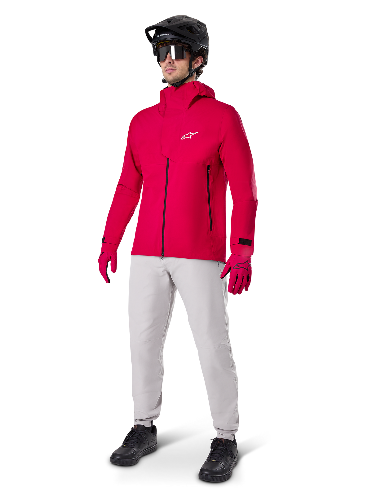 Ciclista masculino com o Casaco Impermeável Alpinestars A-Dura Elite em Vermelho Fluo, calças técnicas cinzento claro, luvas vermelhas, capacete de BTT preto com óculos e sapatos pretos, equipamento completo de alto desempenho para mountain biking