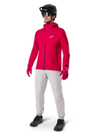 Ciclista masculino com o Casaco Impermeável Alpinestars A-Dura Elite em Vermelho Fluo, calças técnicas cinzento claro, luvas vermelhas, capacete de BTT preto com óculos e sapatos pretos, equipamento completo de alto desempenho para mountain biking