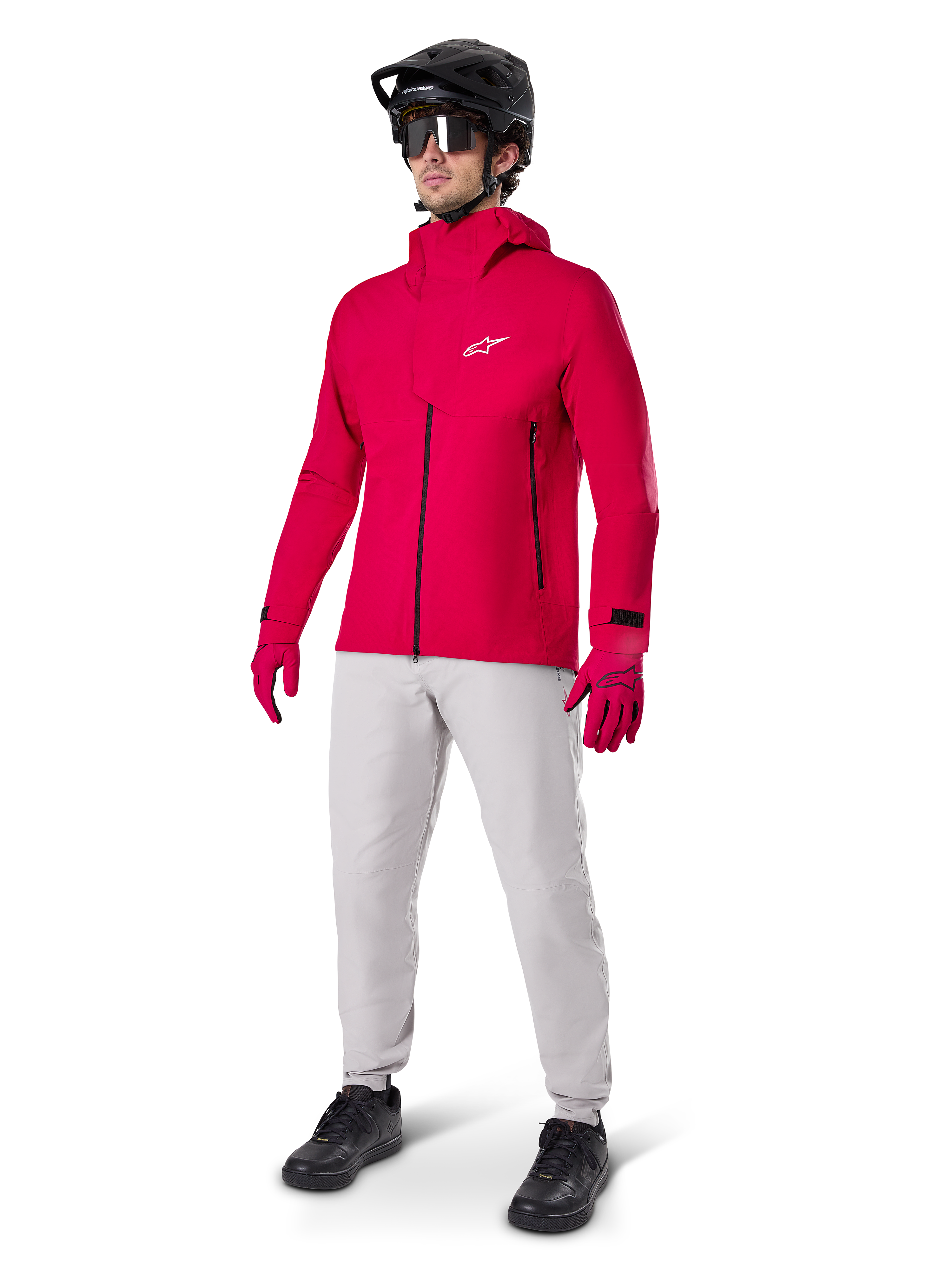 Ciclista masculino com o Casaco Impermeável Alpinestars A-Dura Elite em Vermelho Fluo, calças técnicas cinzento claro, luvas vermelhas, capacete de BTT preto com óculos e sapatos pretos, equipamento completo de alto desempenho para mountain biking