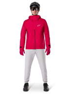 Casaco Impermeável Alpinestars A-Dura Elite, Vermelho Fluo, casaco técnico de BTT usado com um capacete de BTT preto, óculos de sol, luvas vermelhas a condizer e calças cinzentas claras, vista de corpo inteiro de equipamento de ciclismo para todas as condições meteorológicas