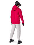Casaco Impermeável Alpinestars A-Dura Elite, Vermelho Fluo, vista traseira de três quartos, usado com calças cinzento claro e capacete de BTT preto, inclui capuz e detalhes refletores, casaco de chuva de ciclismo de alto desempenho para trilhos