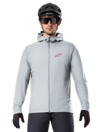 Alpinestars A-Dura Thermal Jacket em Titanium, casaco de ciclismo cinzento claro com capuz e logótipo de estrela vermelho no peito, usado por um ciclista com capacete de BTT preto, óculos de sol e luvas, equipamento térmico para mountain biking