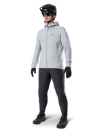 Casaco Térmico Alpinestars A-Dura em Titanium, casaco de ciclismo cinzento claro com capuz e um logótipo Alpinestars vermelho no peito, usado por um homem com capacete de BTT preto, óculos de sol, calças cinzento-escuras e luvas pretas, equipamento profissional de BTT