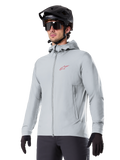 Alpinestars A-Dura Thermal Jacket, casaco térmico de ciclismo em cinzento claro, design com capuz e um pequeno logótipo vermelho, usado com capacete e luvas pretas, vestuário técnico de BTT para condução em tempo frio