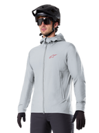 Alpinestars A-Dura Thermal Jacket, casaco térmico de ciclismo em cinzento claro, design com capuz e um pequeno logótipo vermelho, usado com capacete e luvas pretas, vestuário técnico de BTT para condução em tempo frio