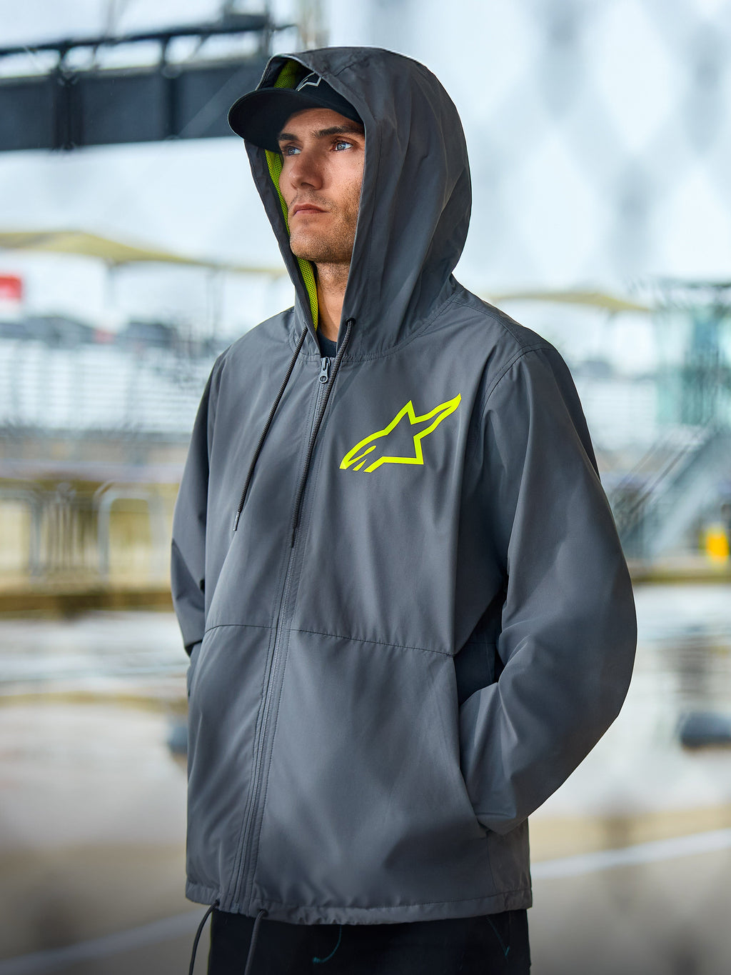 Casaco Corta-vento Alpinestars Speeder Chromium, casaco lifestyle cinzento carvão com detalhes em amarelo de alta visibilidade, design com capuz com forro interior e logótipo no peito amarelos, streetwear casual de desportos motorizados usado por um modelo num ambiente exterior