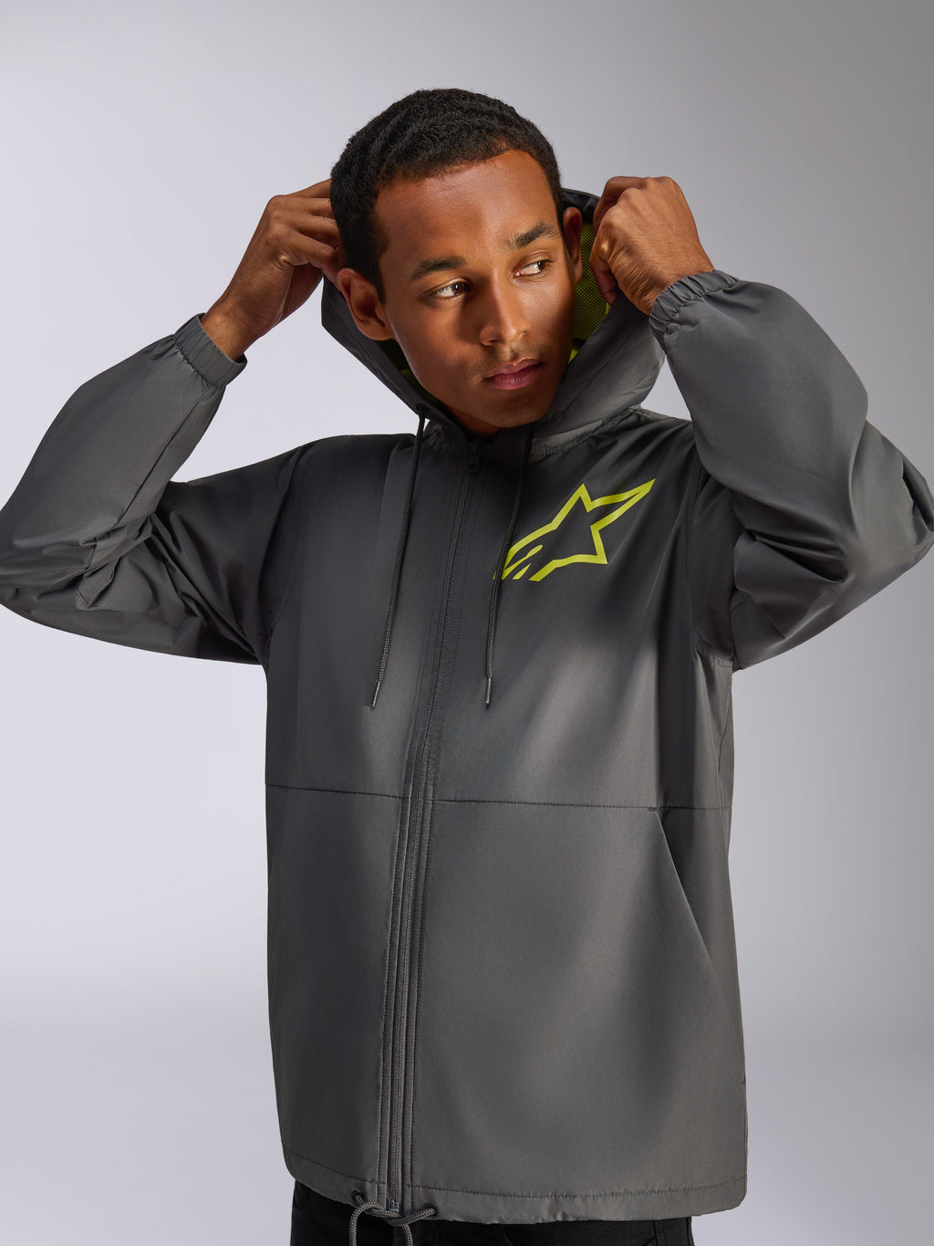 Casaco Corta-vento Alpinestars Speeder Chromium, cinzento carvão e amarelo de alta visibilidade, modelo masculino a usar casaco corta-vento com capuz e cordão, logótipo Alpinestars amarelo de alta visibilidade no peito e forro do capuz amarelo, streetwear casual inspirado no desporto motorizado