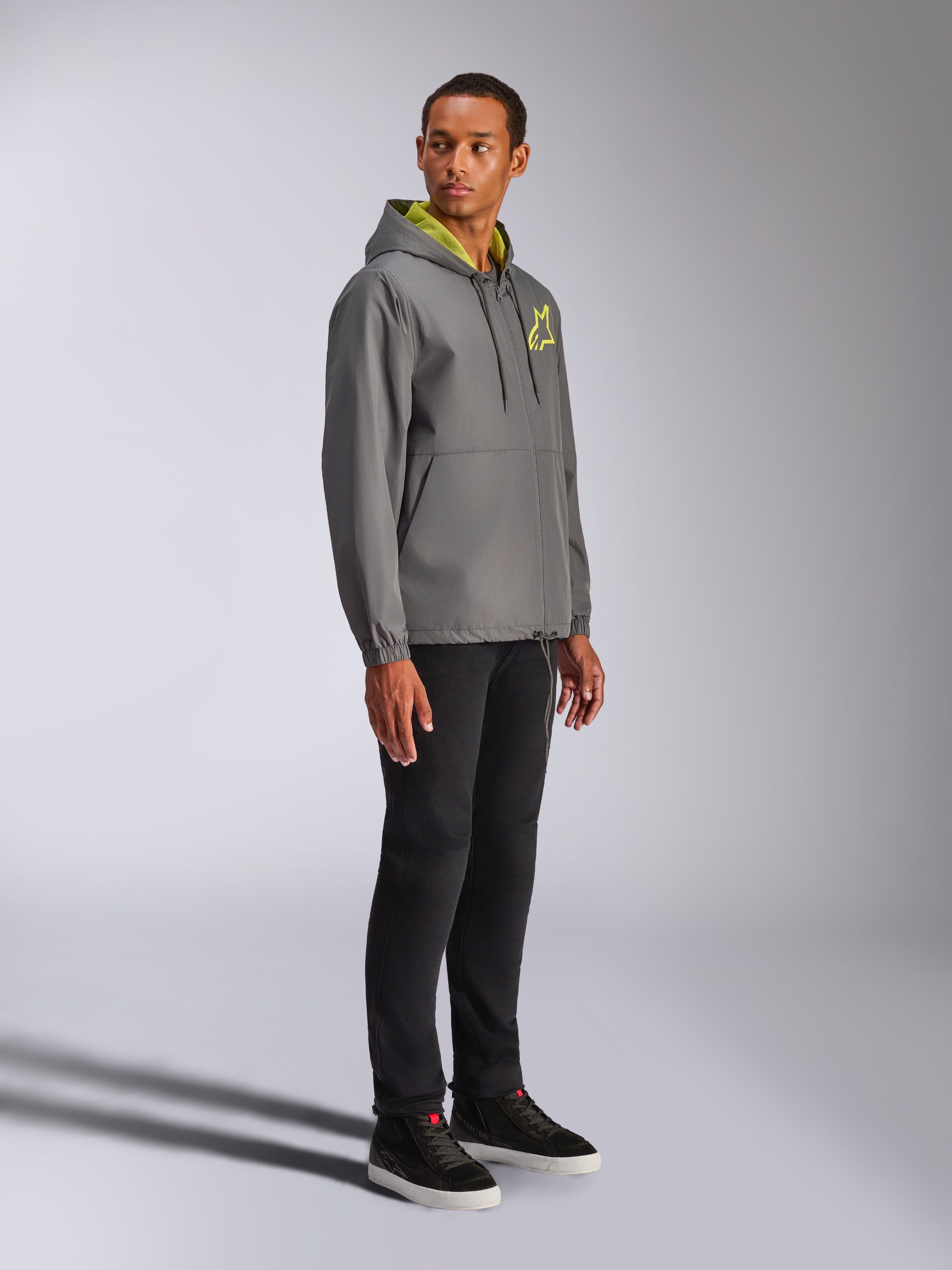 Alpinestars Speeder Chromium Windbreak Jacket, casaco corta-vento cinzento carvão com detalhes em amarelo néon, modelo masculino a usar casaco com capuz, logótipo de estrela amarela no peito, combinado com calças pretas e sapatilhas pretas, streetwear casual inspirado no desporto motorizado