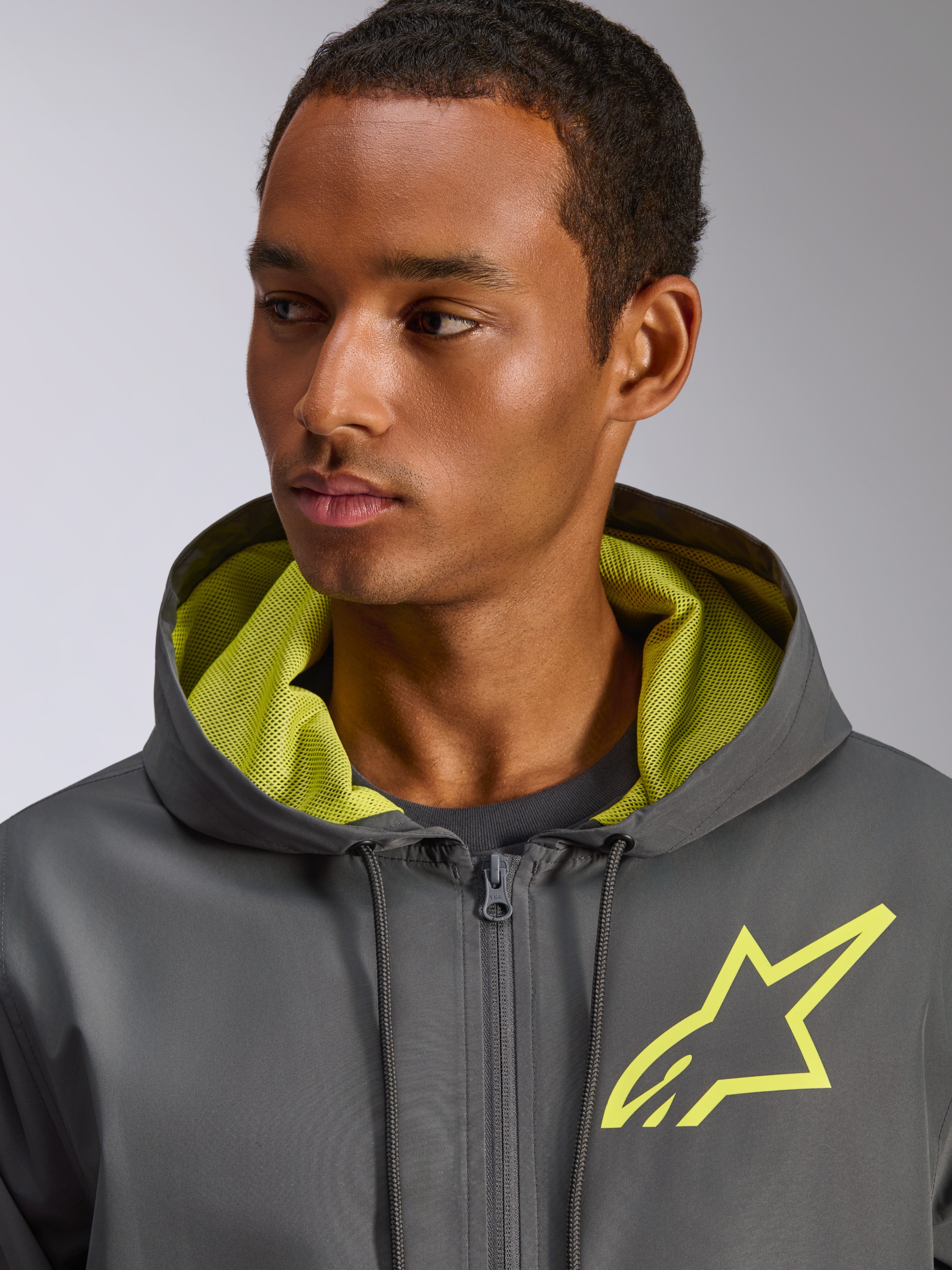 Casaco Corta-vento Alpinestars Speeder Chromium, cinzento antracite com detalhes em amarelo de alta visibilidade, logótipo de estrela amarela no peito, forro do capuz em malha a condizer, corta-vento casual leve
