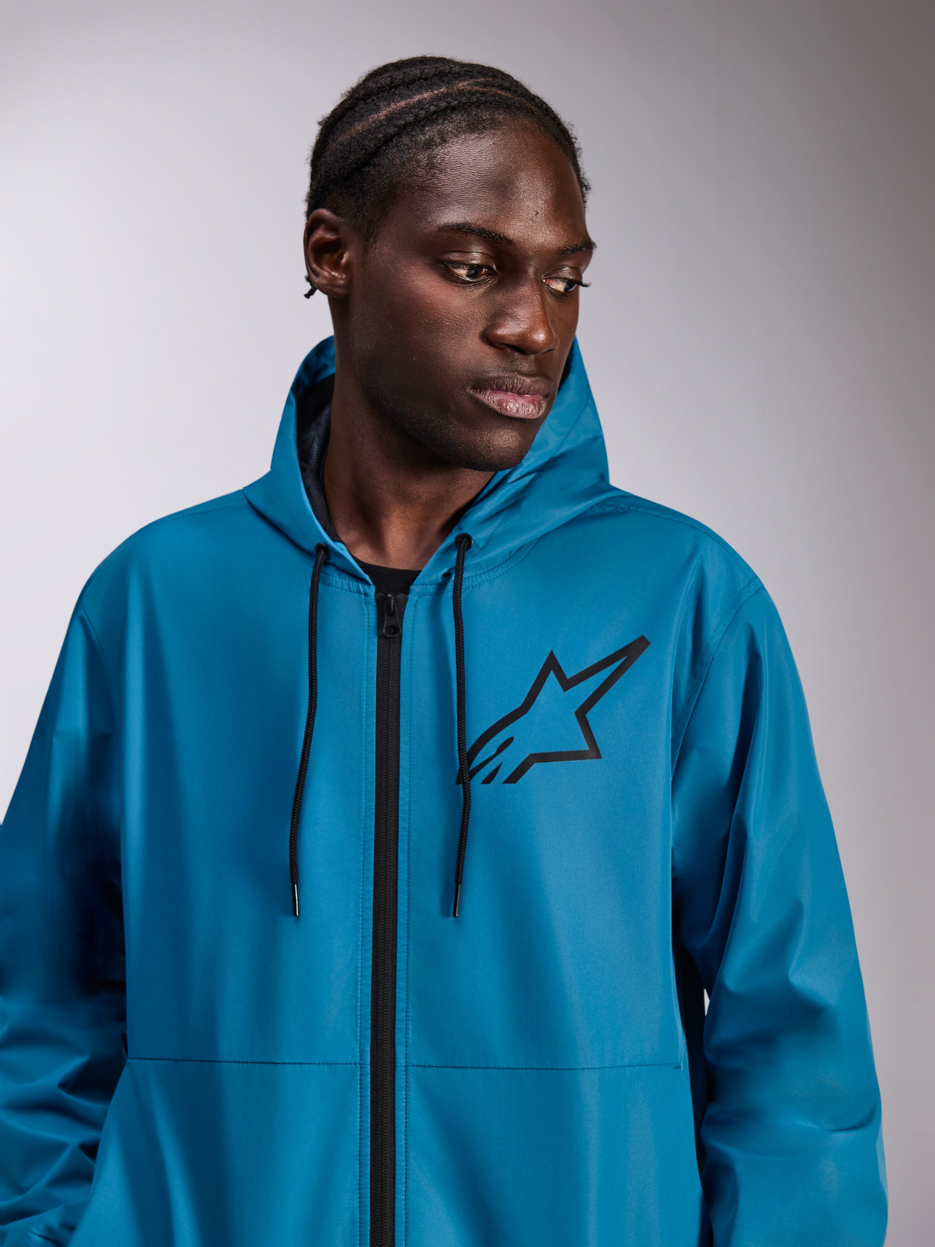 Modelo masculino a usar o casaco corta-vento Alpinestars Speeder Chromium, azul, corta-vento leve com capuz, logótipo Astars preto no peito, fecho de correr frontal e cordões pretos, streetwear casual inspirado no desporto motorizado