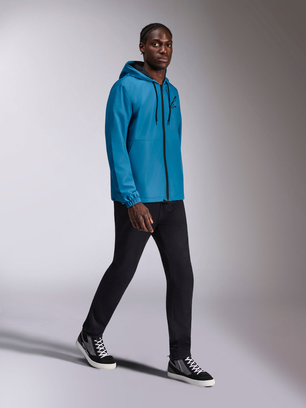 Alpinestars Speeder Chromium Windbreak Jacket, corta-vento azul com capuz usado por um modelo masculino, apresenta fecho de correr frontal preto e logótipo no peito, combinado com calças pretas e sapatilhas de cano alto, vestuário desportivo casual inspirado no desporto motorizado