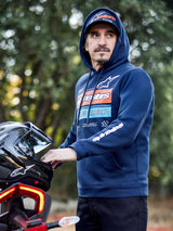Alpinestars Harken Hoodie, azul marinho, hoodie de lã com gráfico em degradê no peito e logótipo Alpinestars branco na manga, vestido por um homem a segurar um capacete de mota, streetwear casual inspirado no desporto motorizado