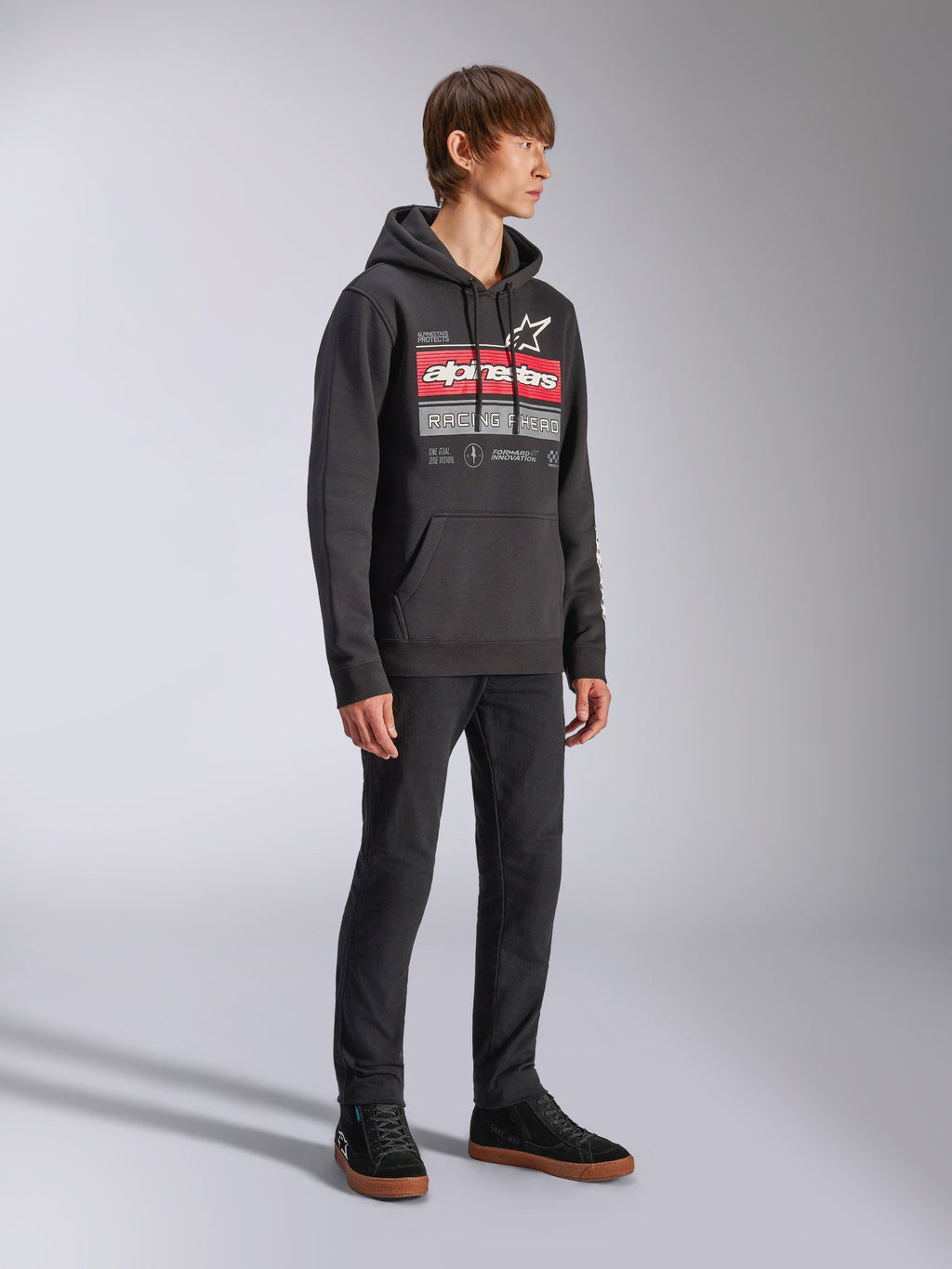 Alpinestars Harken Hoodie, hoodie de lã preto, com um grafismo grande no peito apresentando a marca Alpinestars a vermelho e o logótipo da estrela, bolso canguru, vestido por um modelo com calças pretas e sapatilhas, streetwear casual de desportos motorizados