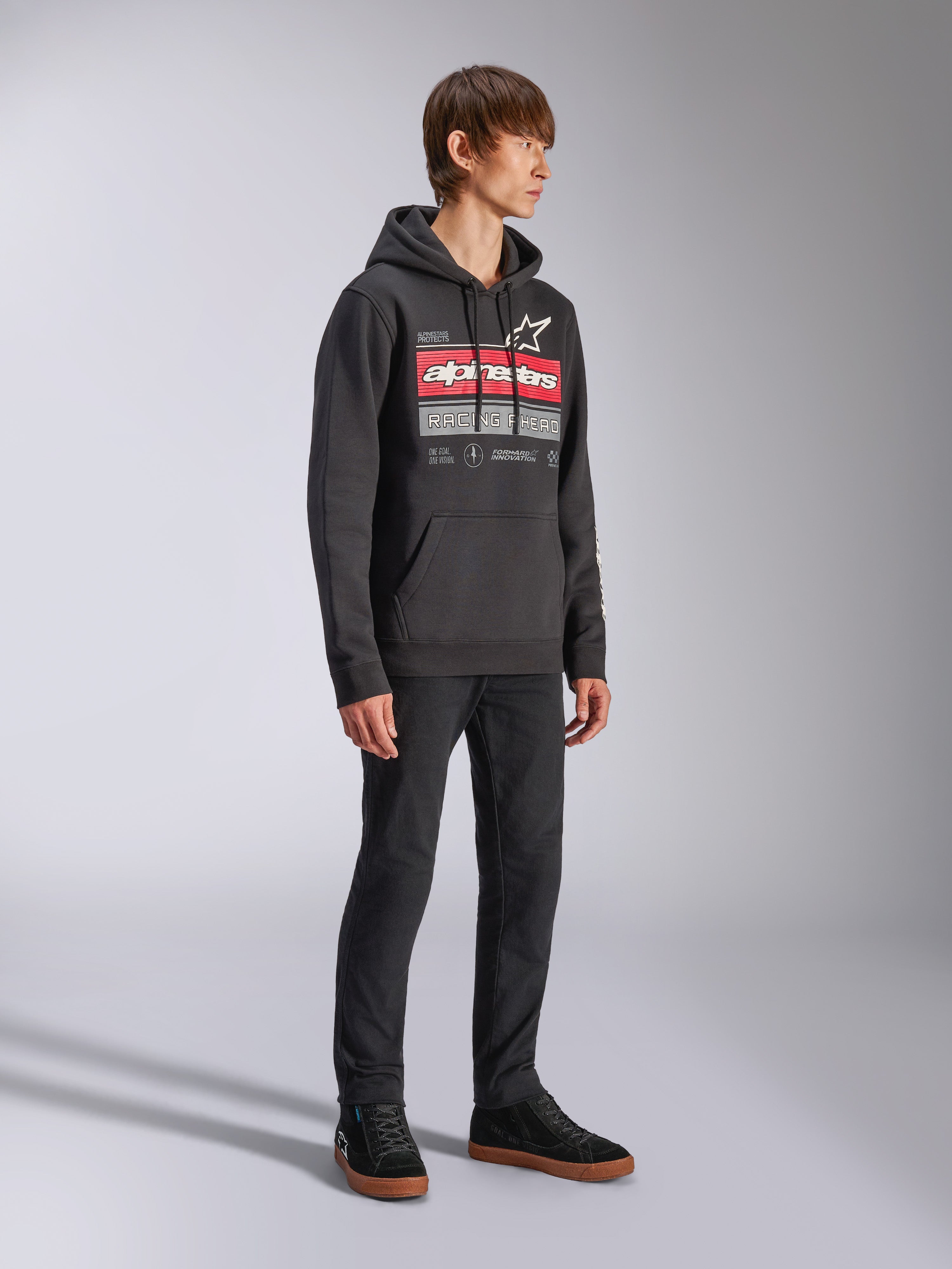 Alpinestars Harken Hoodie, hoodie de lã preto, com um grafismo grande no peito apresentando a marca Alpinestars a vermelho e o logótipo da estrela, bolso canguru, vestido por um modelo com calças pretas e sapatilhas, streetwear casual de desportos motorizados