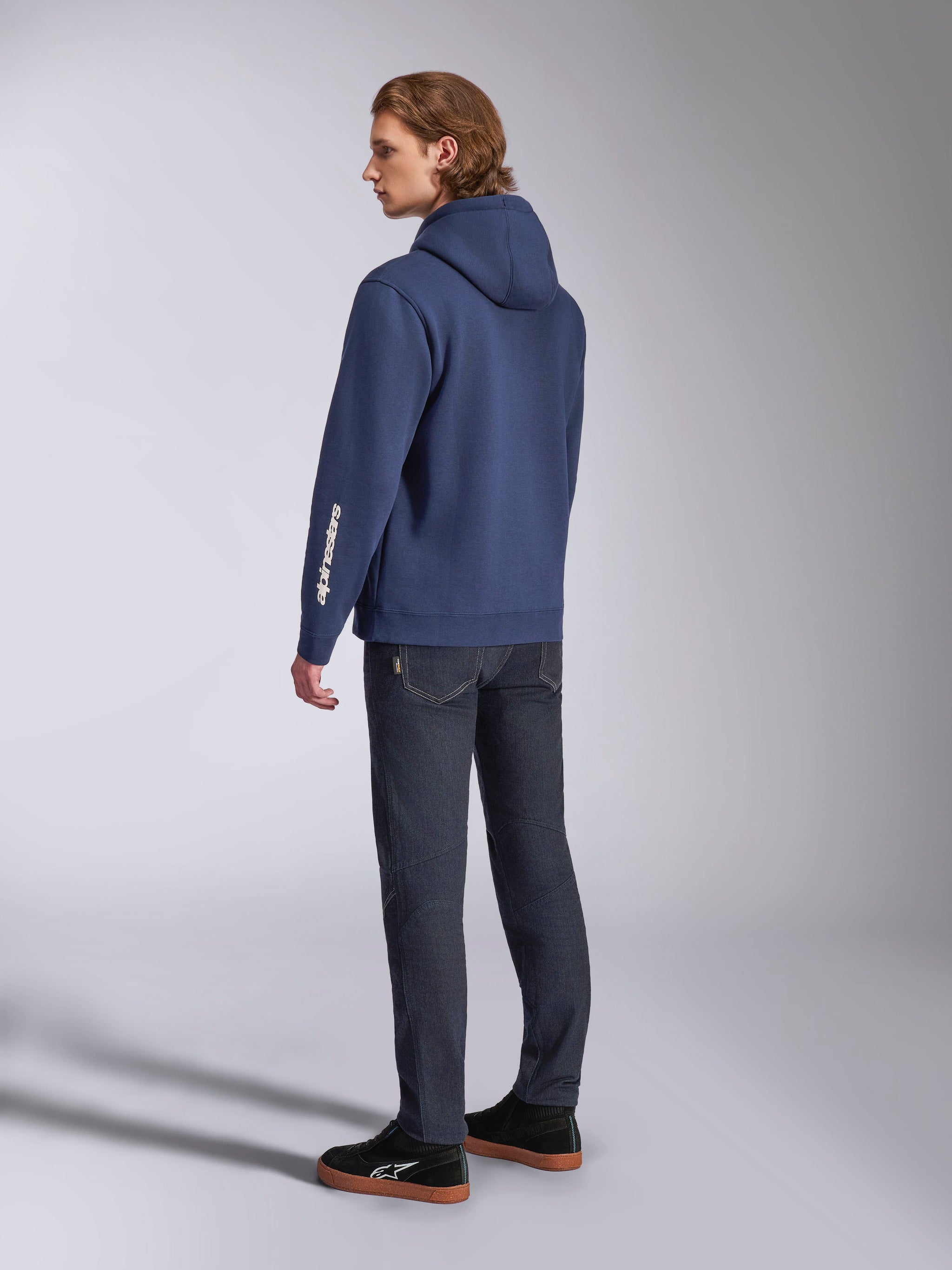 Alpinestars Harken Hoodie em azul-marinho, vista traseira de modelo masculino, hoodie casual em lã com logótipo de texto branco na manga, combinado com calças de ganga escuras e sapatilhas pretas, streetwear moderno inspirado no desporto motorizado