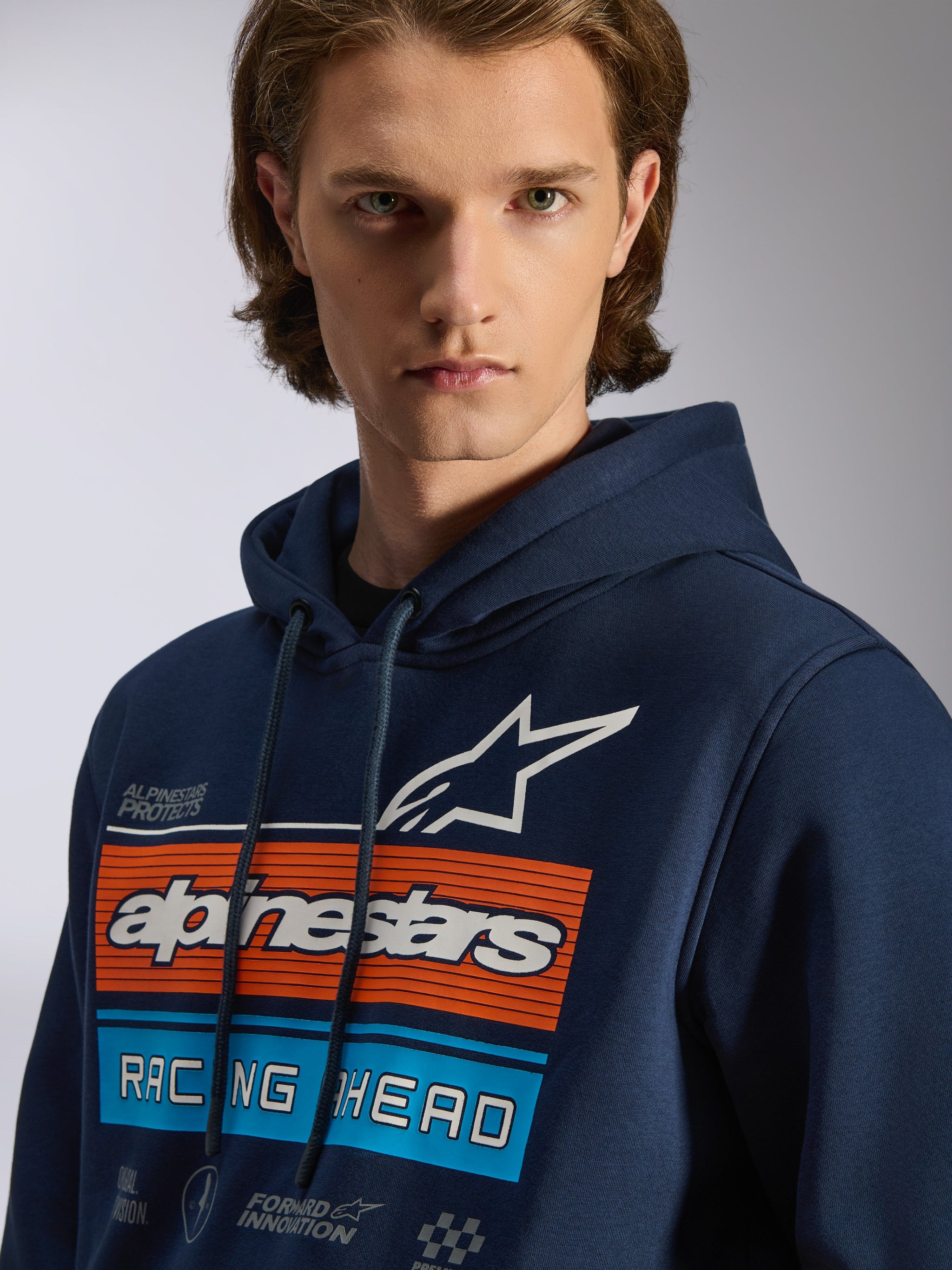 Alpinestars Harken Hoodie, hoodie pullover azul marinho, grafismo no peito com barras horizontais cor de laranja e azul claro, logótipo de estrela branco, texto 'Racing Ahead', vestuário casual de estilo motorsport