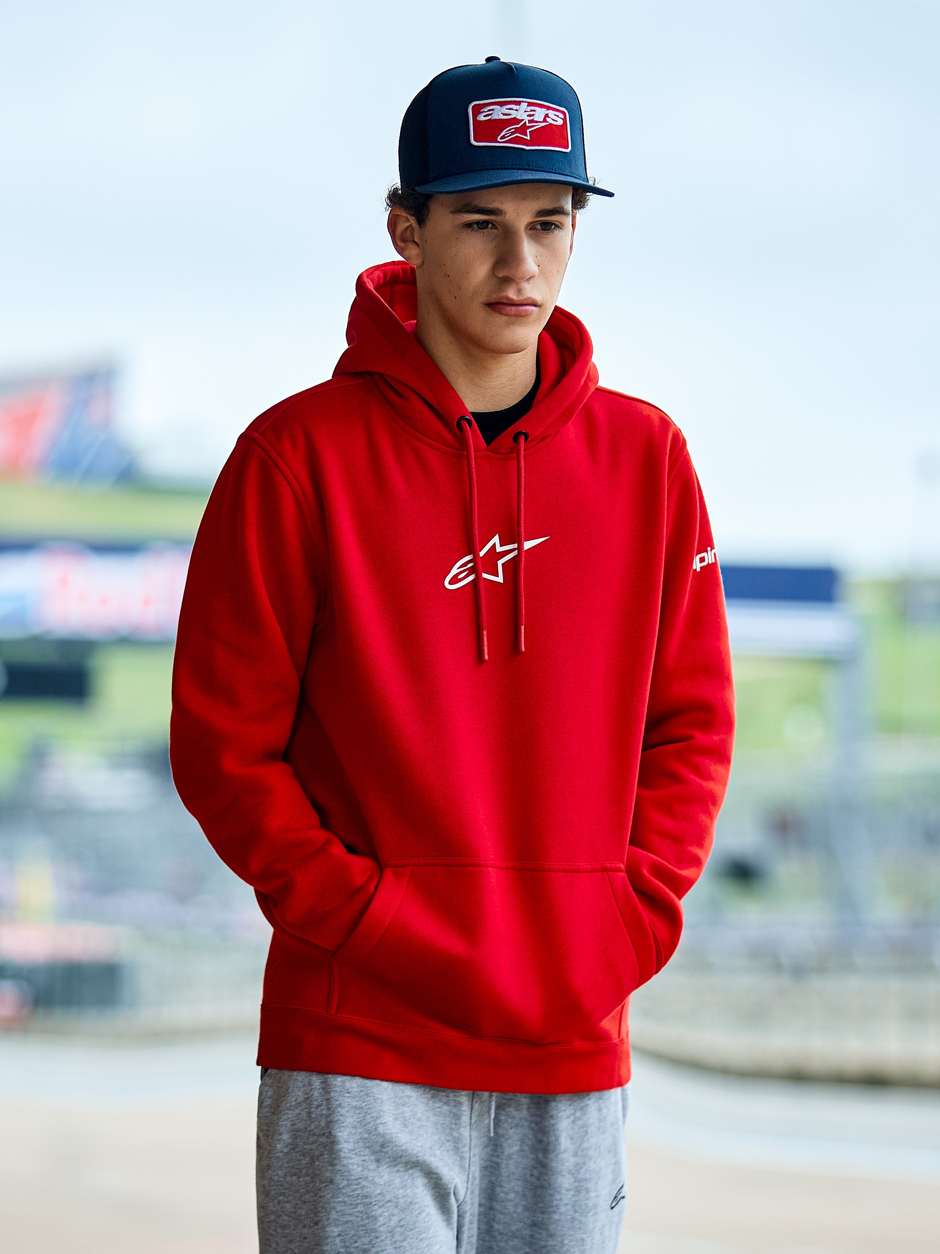 Alpinestars Frontal Hoodie, hoodie em lã vermelha, com logótipo Astars branco no peito, bolso canguru e capuz com cordão, usado por um homem com boné azul, streetwear casual de motorsport