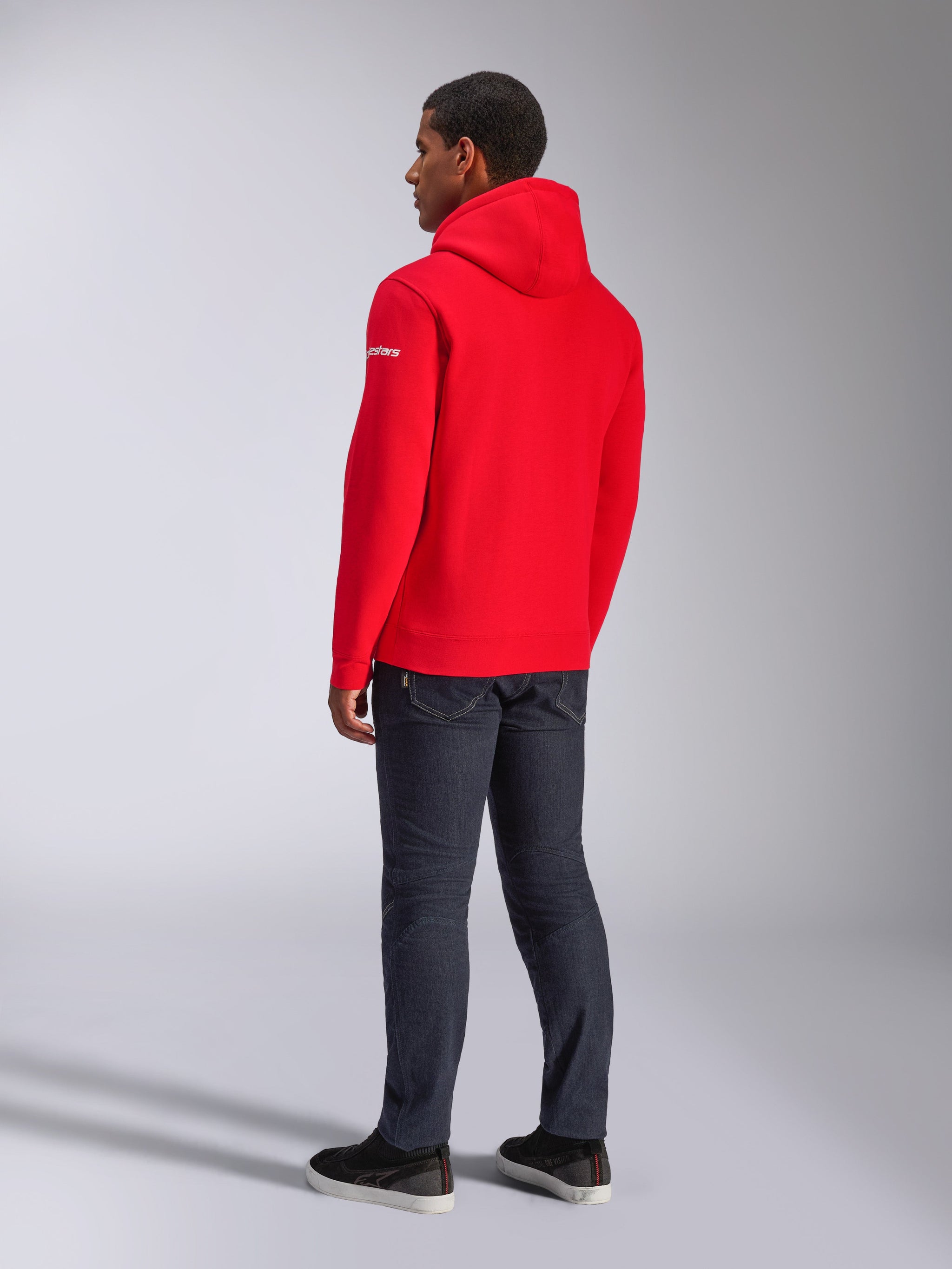 Alpinestars Frontal Hoodie, hoodie vermelho de lã com capuz, vista traseira que mostra o logótipo branco na manga, usado com calças de ganga escuras e sapatos pretos, streetwear casual de desporto motorizado