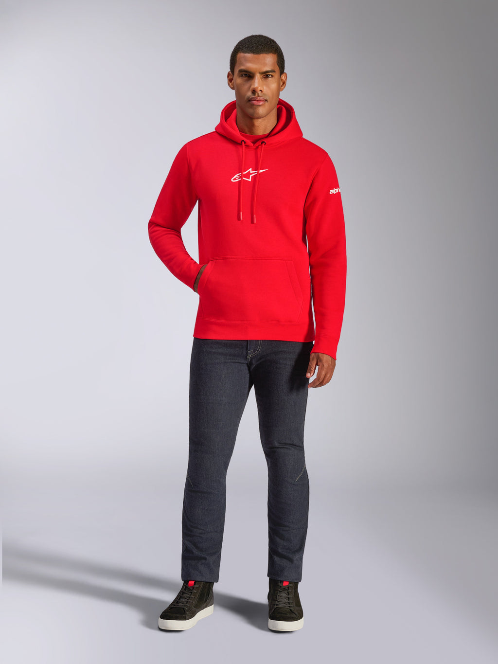 Modelo masculino a usar o Alpinestars Frontal Hoodie em vermelho, hoodie de lã com logótipo Astars branco no peito, bolso canguru e capuz com cordão, combinado com calças de ganga cinzento-escuras e sapatilhas de cano alto pretas, estilo urbano casual de desportos motorizados
