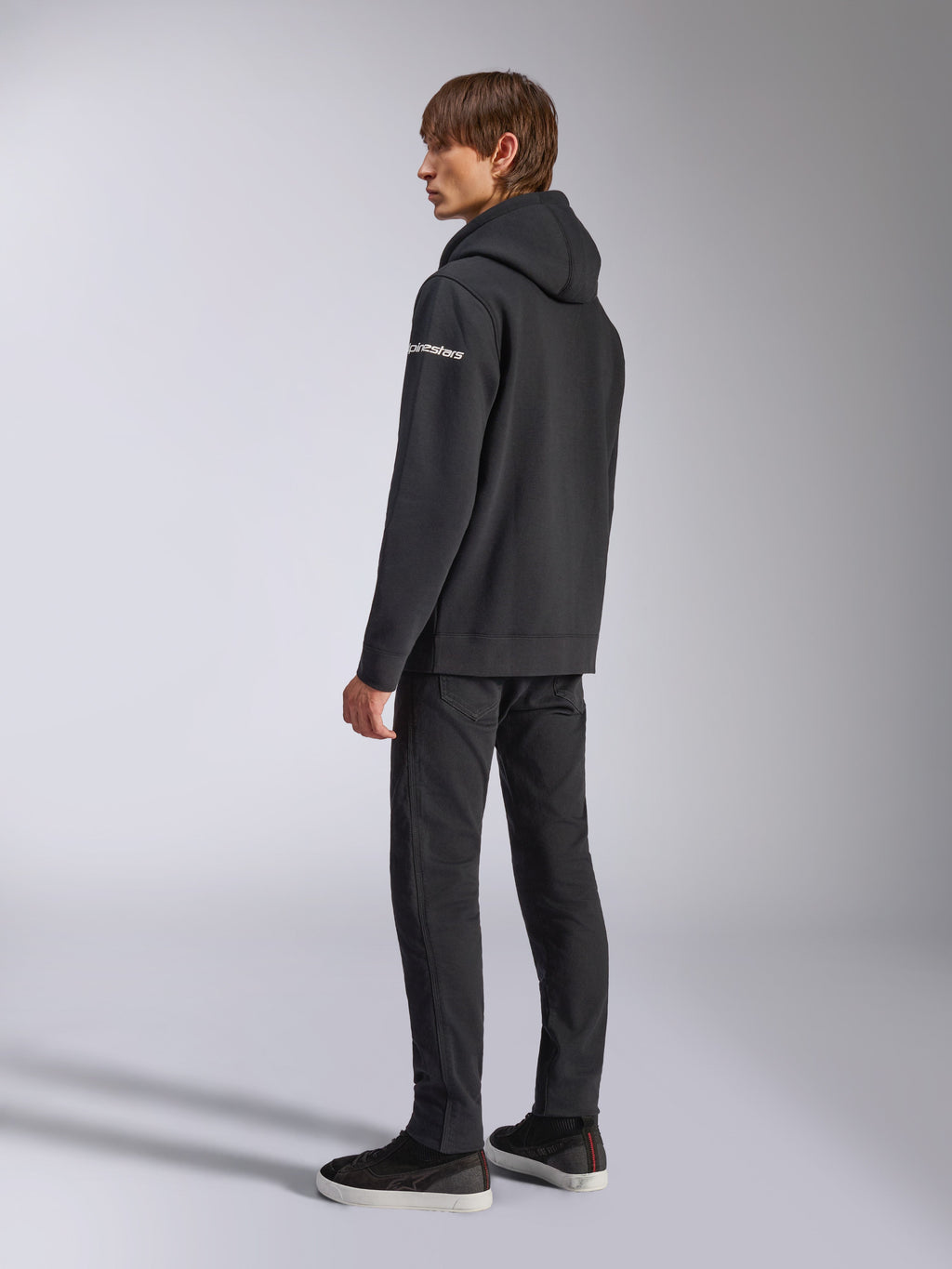 Alpinestars Frontal Hoodie, streetwear casual, preto, vista lateral traseira mostrando o logótipo nominal branco na manga, usado com calças pretas e sapatilhas, design moderno inspirado no desporto motorizado