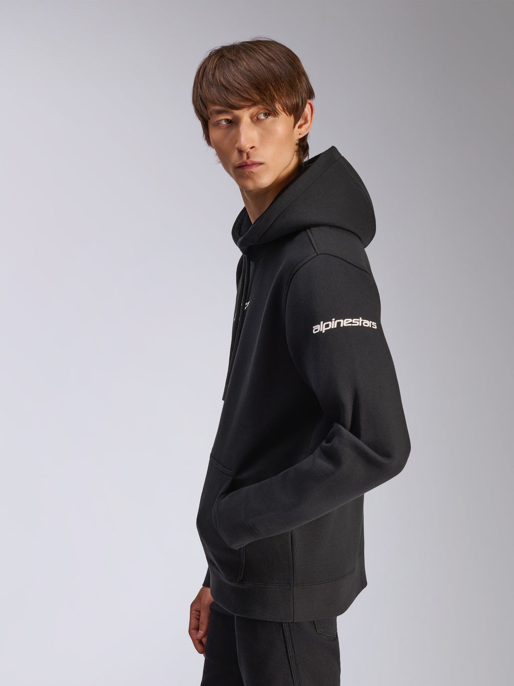 Alpinestars Frontal Hoodie, hoodie de lã preto, modelo visto de perfil lateral, logótipo Alpinestars branco na manga, bolso canguru, streetwear casual inspirado no desporto motorizado