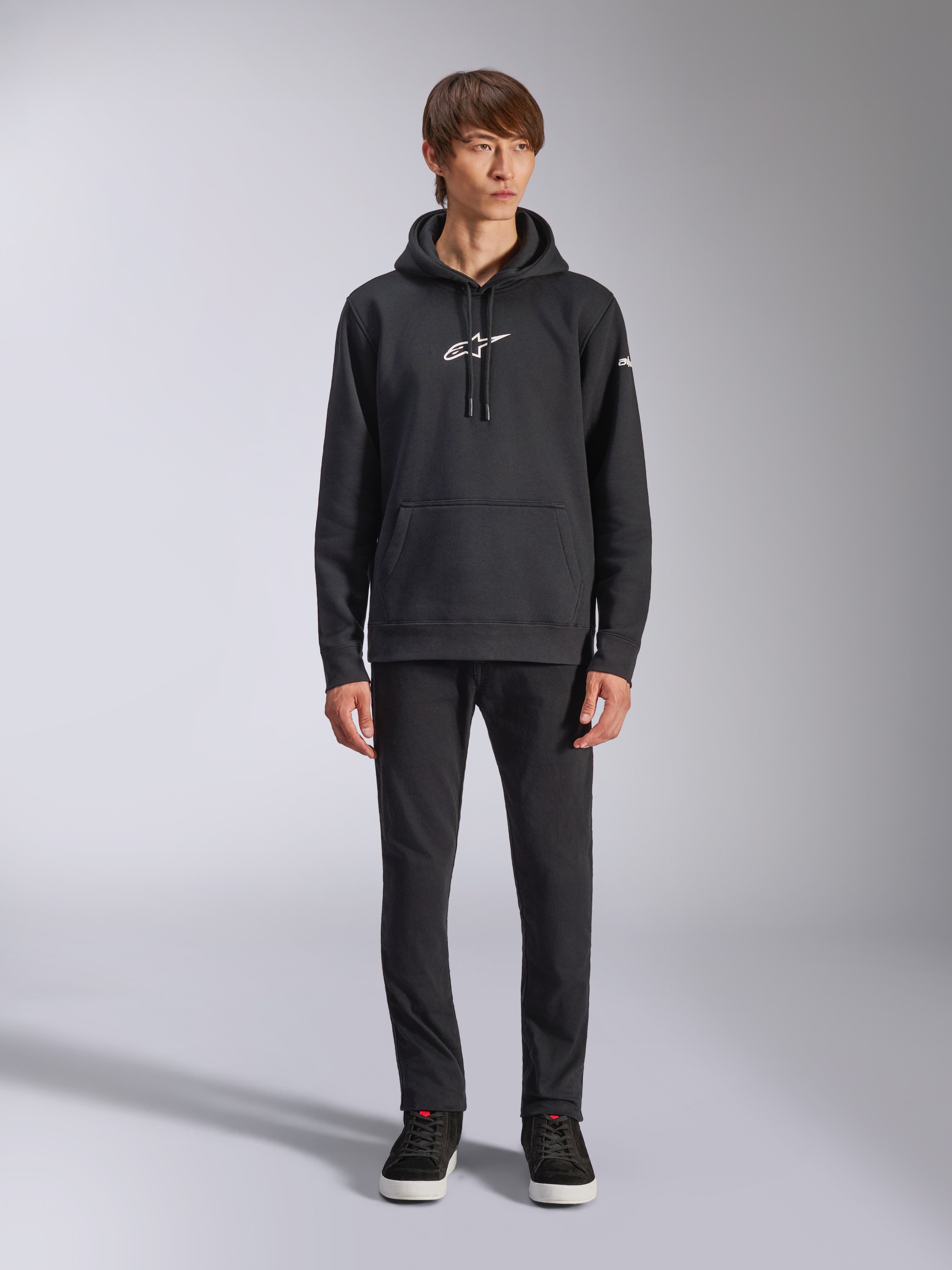 Alpinestars Frontal Hoodie, hoodie de lã preto, logótipo Alpinestars branco no peito e na manga, bolso canguru, capuz com cordão, usado com calças pretas, design casual inspirado no desporto motorizado