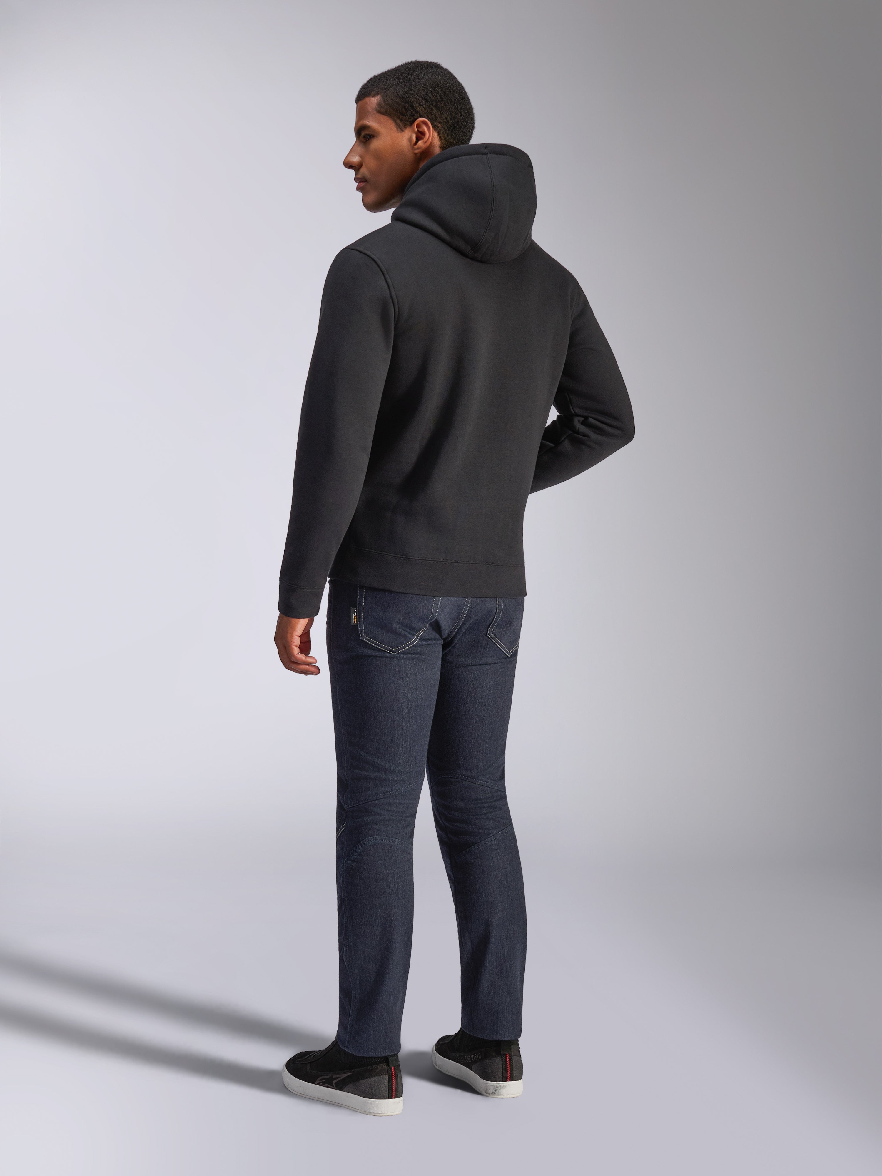 Alpinestars Blaze V3 Hoodie, vista traseira de um homem a usar a camisola preta com capuz, combinada com calças de ganga escuras e sapatilhas pretas, streetwear casual inspirado no desporto motorizado