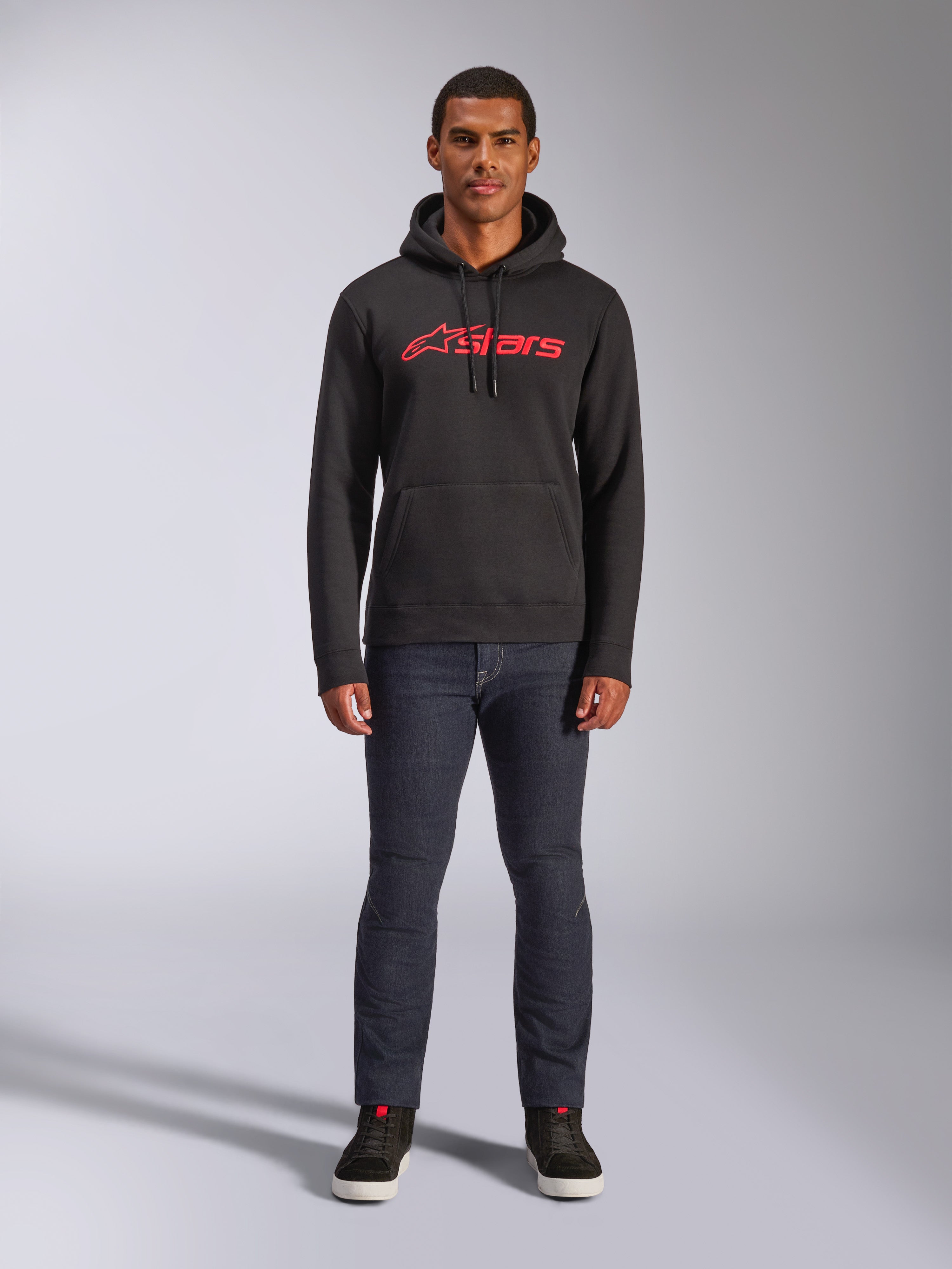 Alpinestars Blaze V3 Hoodie, camisola com capuz de estilo streetwear casual, preta com logótipo Astars vermelho, vista frontal, usada por um modelo com calças de ganga escuras e sapatilhas pretas, design moderno inspirado no desporto motorizado