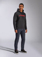 Alpinestars Blaze V3 Hoodie, camisola com capuz preta com logótipo Alpinestars vermelho no peito, usada por um modelo masculino com calças de ganga escuras e sapatilhas pretas, streetwear casual inspirado no desporto motorizado