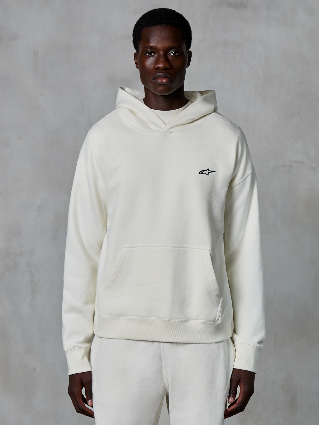 Hoodie Alpinestars Essentials Ageless, off-white, usado por modelo masculino, logótipo pequeno preto no peito, bolso frontal tipo canguru, mangas compridas, streetwear casual inspirado no desporto motorizado