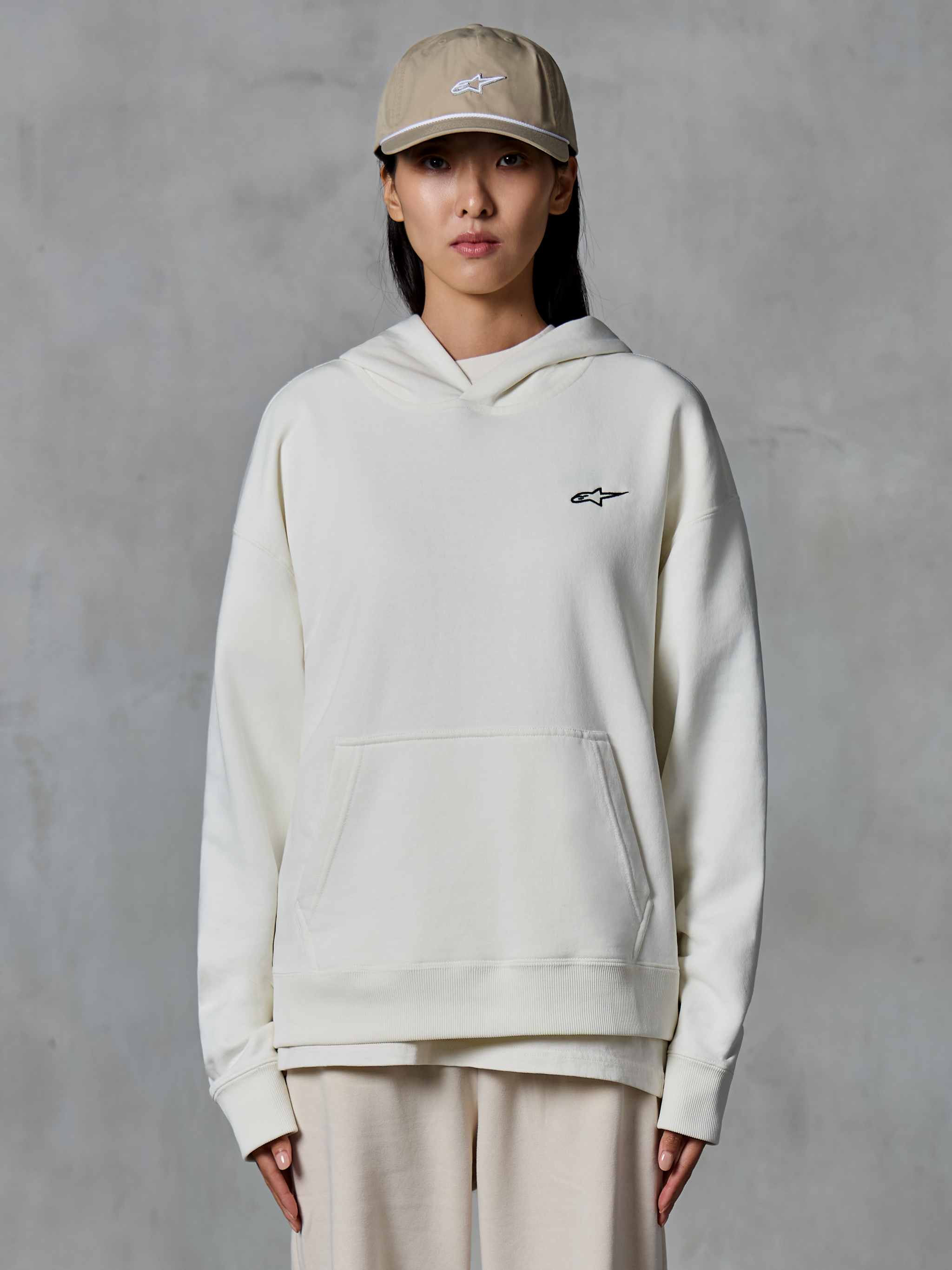 Alpinestars Essentials Ageless Hoodie, hoodie de fleece em off-white, modelo feminina a usar boné bege e calças joggers creme a condizer, pequeno logótipo Alpinestars preto no peito, bolso canguru frontal, corte relaxado, streetwear casual inspirado no desporto motorizado
