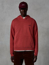 Hoodie Essentials Ageless Alpinestars, streetwear casual, cor vermelho tijolo, com um pequeno logótipo de estrela preto no peito, bolso canguru, acabamento lavado à pedra, usado por um modelo com um boné bordeaux e calças de bombazina castanhas.