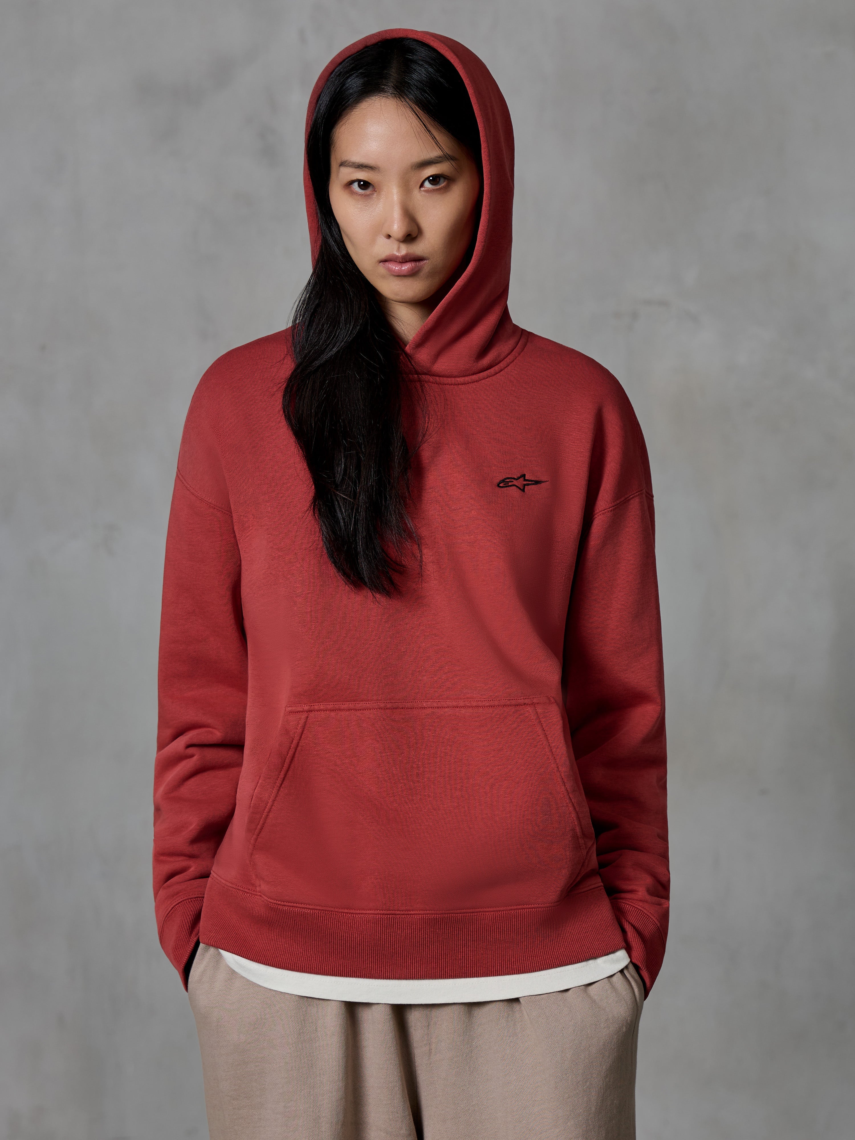 Alpinestars Essentials Ageless Hoodie, sweatshirt pullover em vermelho tijolo, vestida por uma modelo feminina com o capuz colocado, com um pequeno logótipo de estrela bordado a preto no peito, bolso canguru, streetwear casual inspirado no desporto motorizado