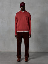 Alpinestars Essentials Ageless Hoodie, hoodie casual de sportswear em vermelho tijolo com acabamento lavado à pedra, apresentando um pequeno logótipo Alpinestars preto no peito e bolso canguru frontal, usado por um modelo com boné bordeaux e calças de bombazina castanhas para um visual streetwear clean.