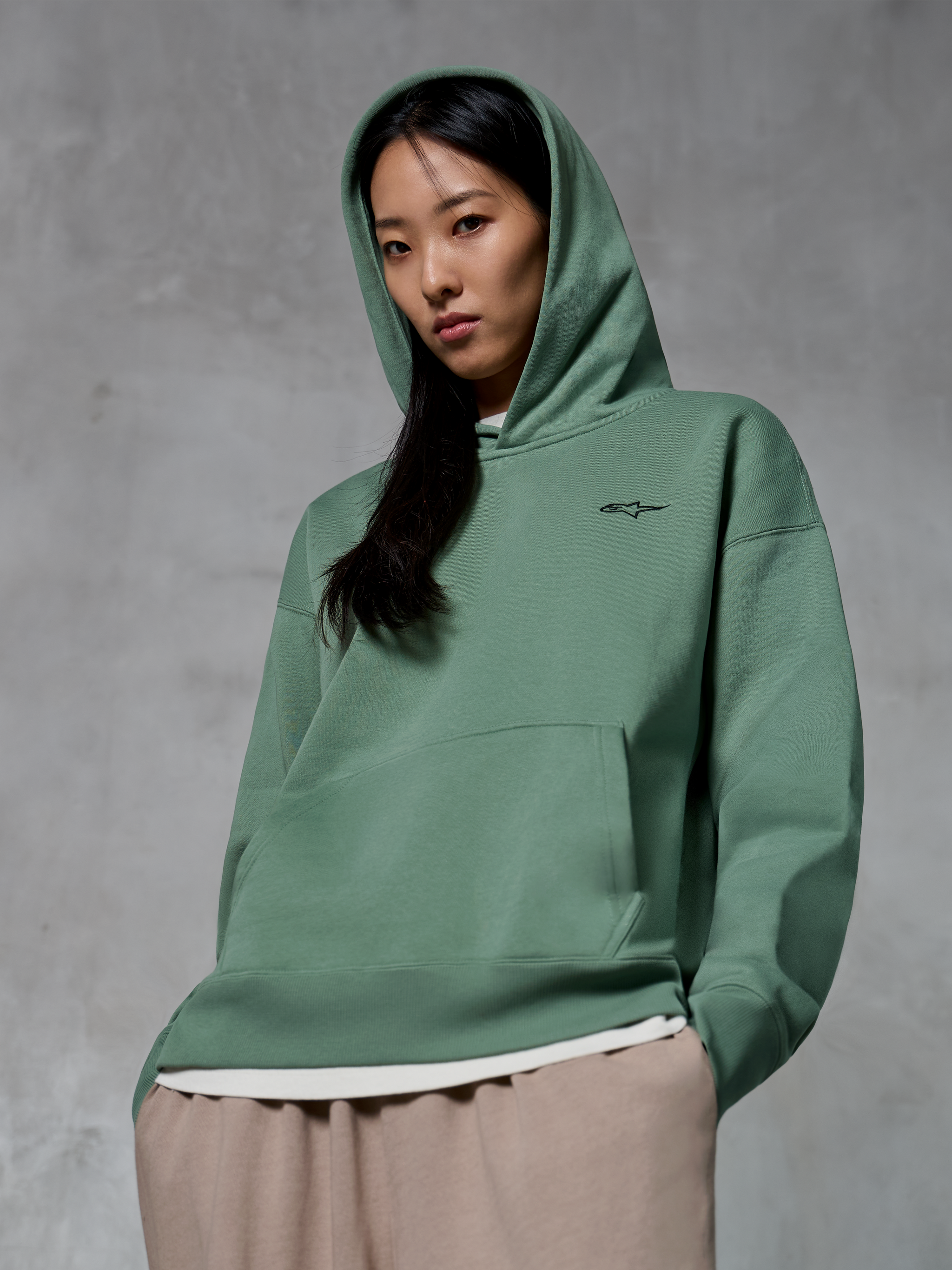 Alpinestars Essentials Ageless Hoodie, casual streetwear, Cactus green with small black logo, hoodie worn with hood up, front kangaroo pocket, stone-washed finish, paired with light tan pants