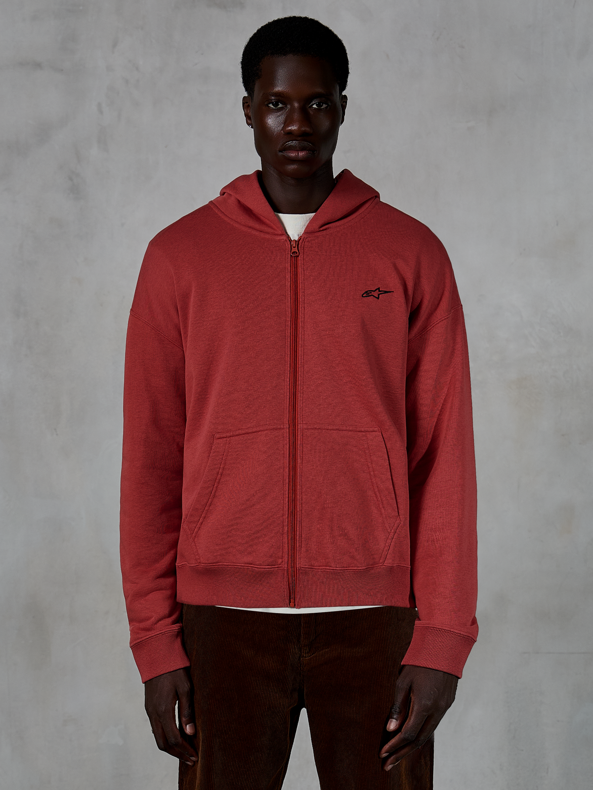 Alpinestars Essentials Ageless Zip Hoodie, sweatshirt com capuz e fecho integral em vermelho tijolo, usada por modelo com calças de bombazina castanhas, com um pequeno logótipo preto no peito, estilo casual streetwear de corte relaxado
