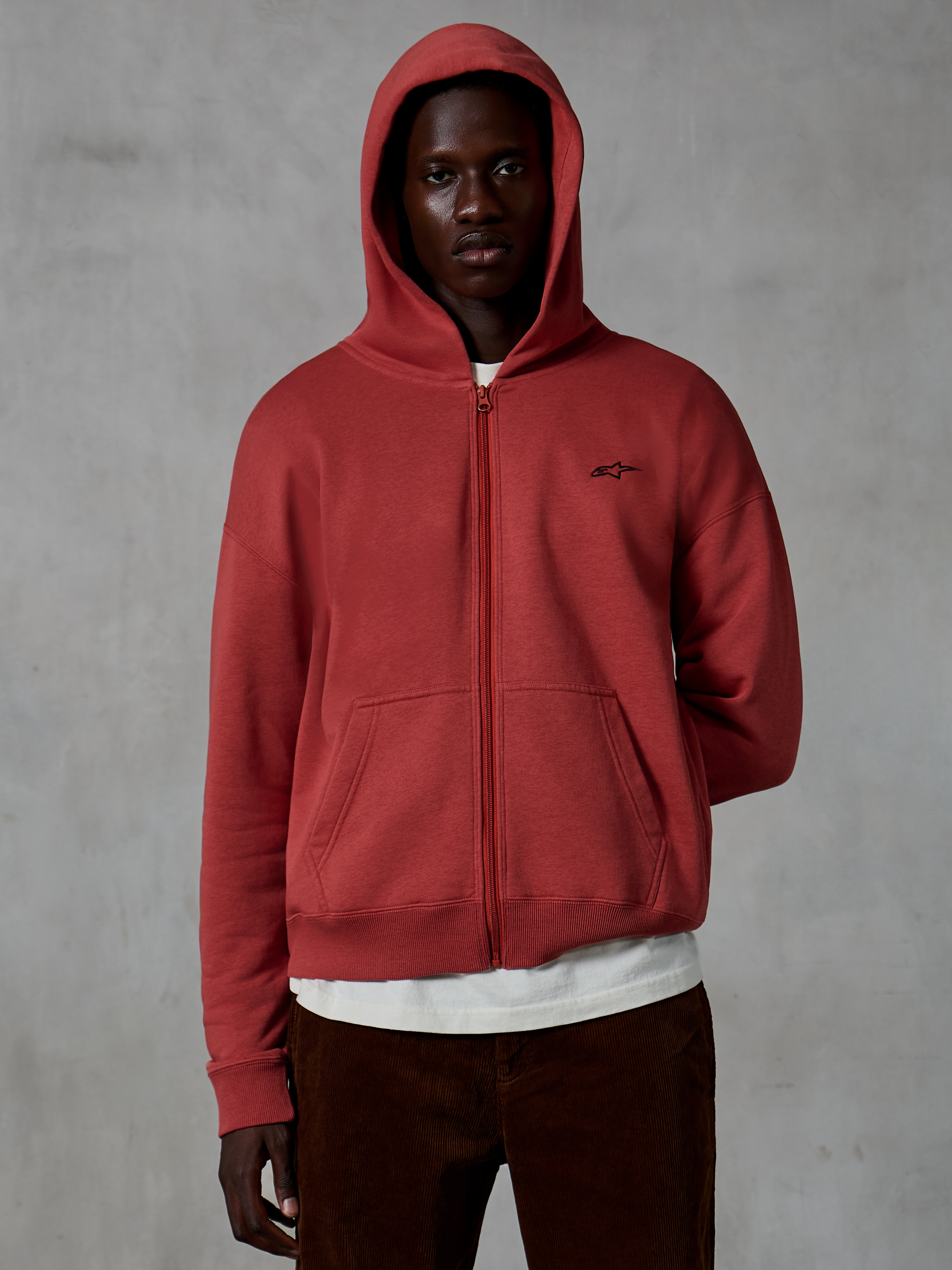 Hoodie Alpinestars Essentials Ageless com fecho, vestuário desportivo casual em vermelho tijolo, design de fecho completo com capuz levantado, com um pequeno logótipo preto no peito, usado por um modelo com t-shirt branca e calças de bombazina castanhas sobre um fundo cinzento.