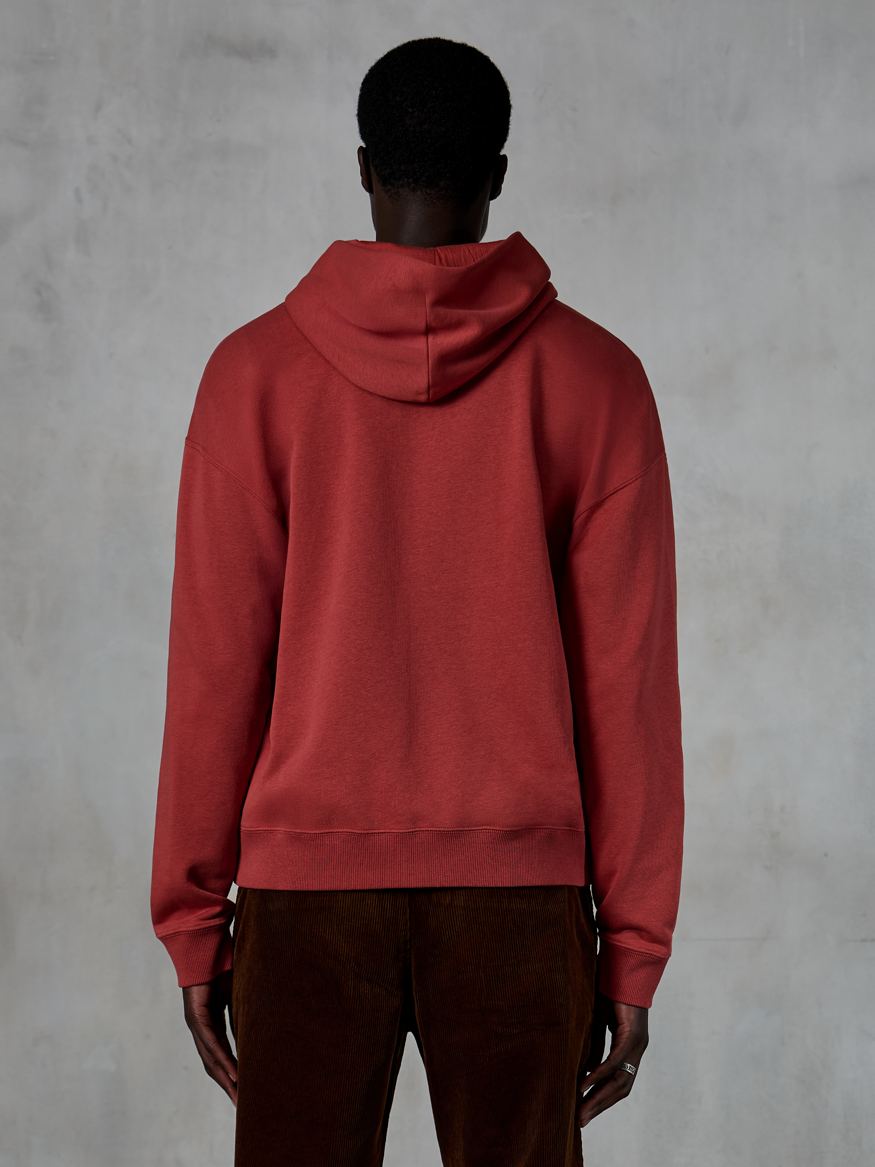 Alpinestars Essentials Ageless Zip Hoodie, vermelho tijolo, vista traseira mostrando o ajuste relaxado e capuz com cordão, usado com calças de bombazina castanhas, streetwear casual inspirado no desporto motorizado
