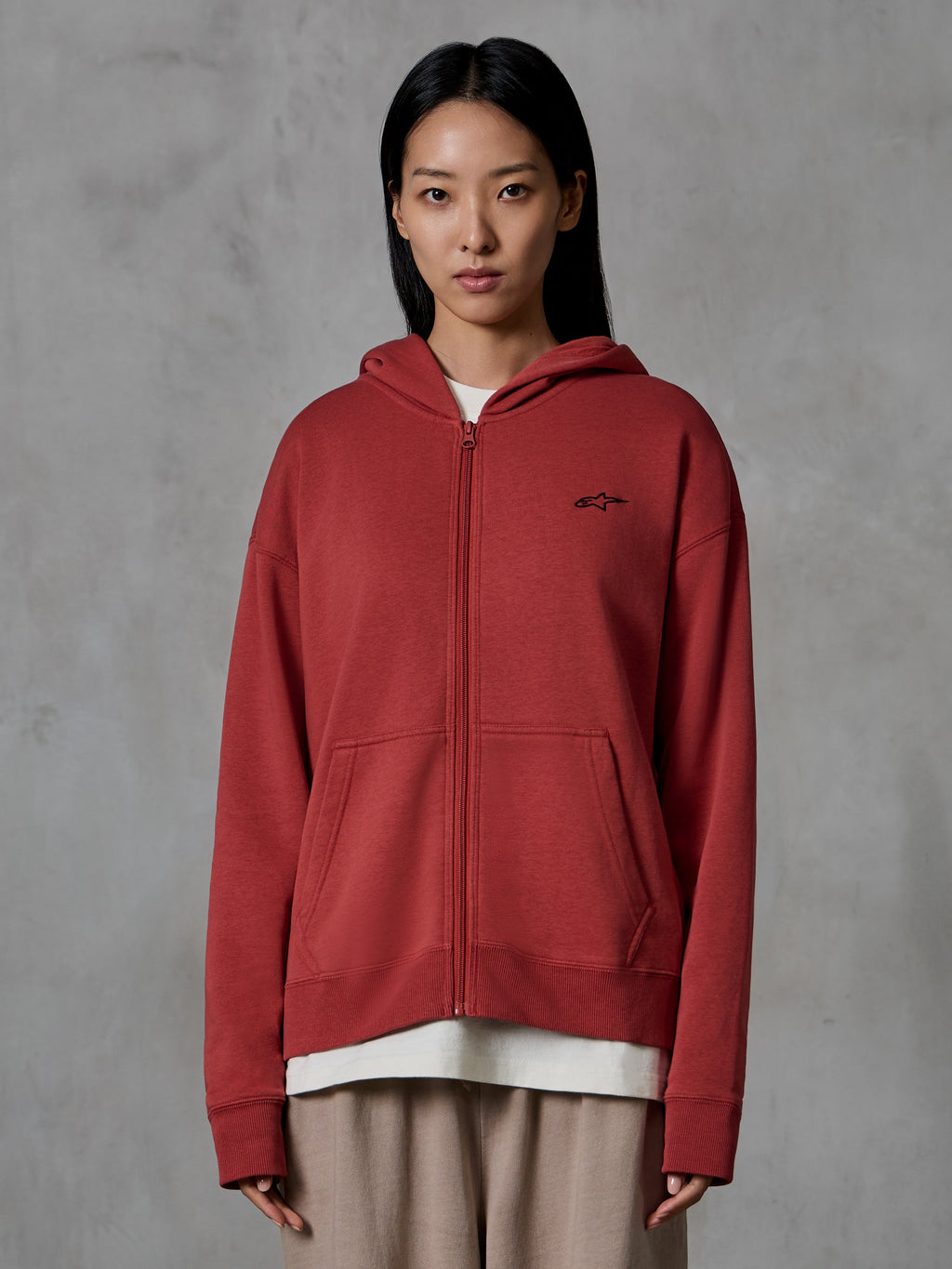 Alpinestars Essentials Ageless Zip Hoodie, vermelho tijolo, streetwear casual, vestido por uma modelo, design com fecho integral e capuz, pequeno logótipo preto da Alpinestars no peito, fleece compacto de corte relaxado