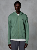 Alpinestars Essentials Ageless Zip Hoodie, hoodie de streetwear casual com fecho, verde Cactus, com um logótipo bordado subtil no peito, bolsos tipo canguru divididos e punhos canelados, vestido por um modelo masculino num fundo cinzento texturizado.