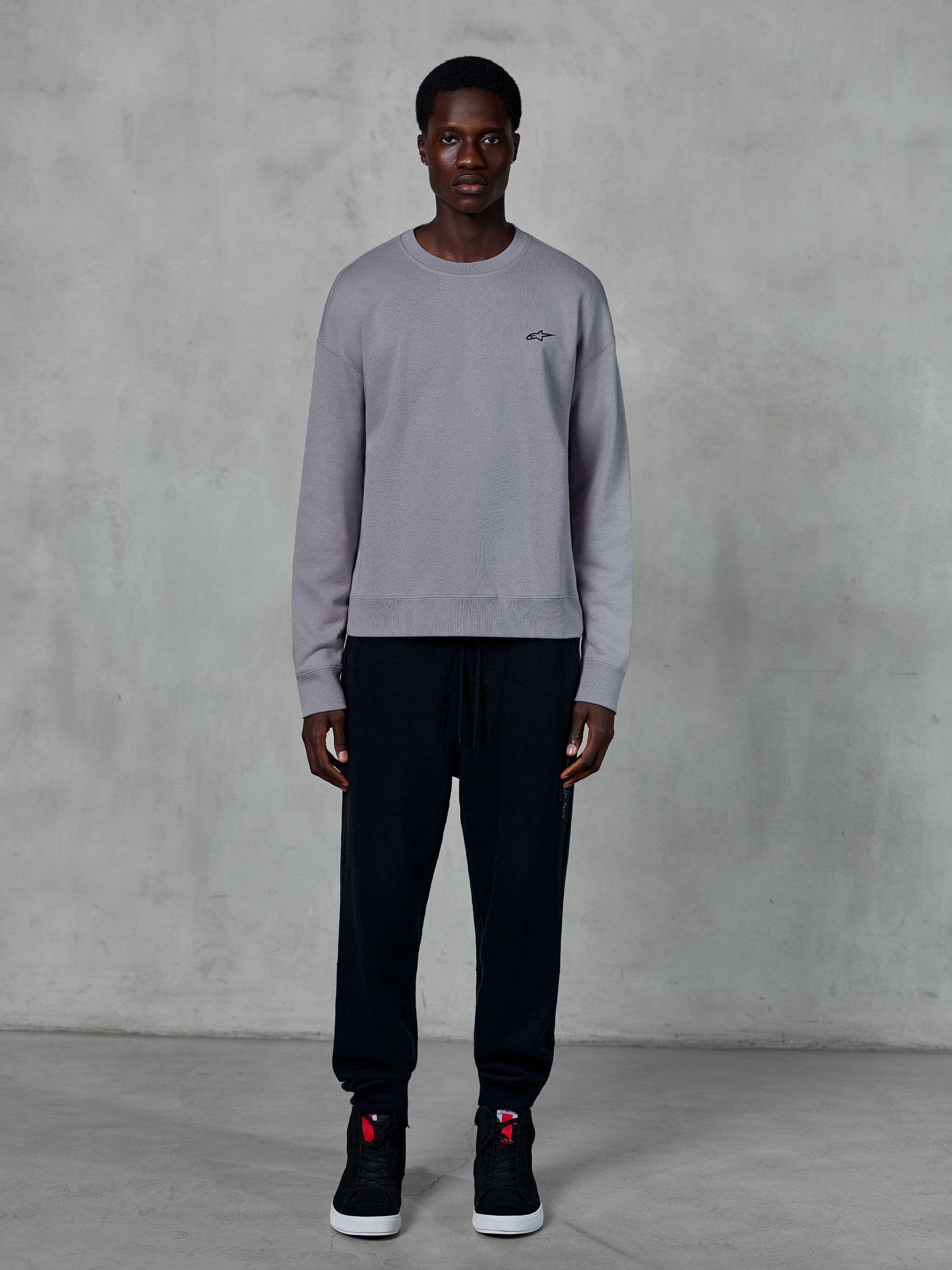 Alpinestars Essentials Ageless Crew, sweatshirt prateada de gola redonda, vestida por um modelo com calças jogger pretas e ténis pretos, com um pequeno logótipo Alpinestars preto no peito, streetwear casual inspirado no desporto motorizado
