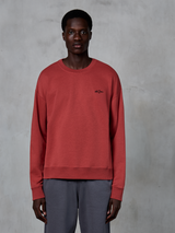 Alpinestars Essentials Ageless Crew, sweatshirt de gola redonda, vermelho tijolo, pequeno logótipo preto no peito, vestida por um modelo com calças cinzentas, fleece macio premium, streetwear casual